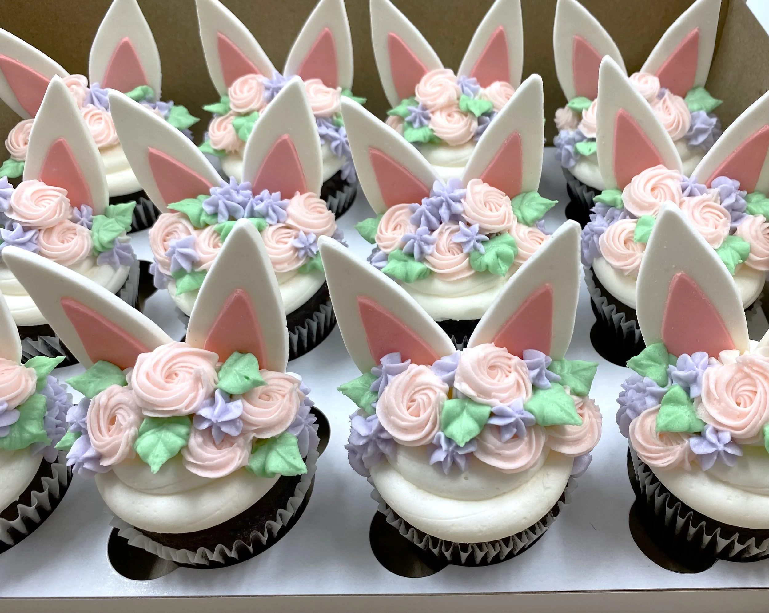 Cupcakes decorated as Easter bunny faces with white and pink bunny ears, pink and purple flowers, and green leaves.