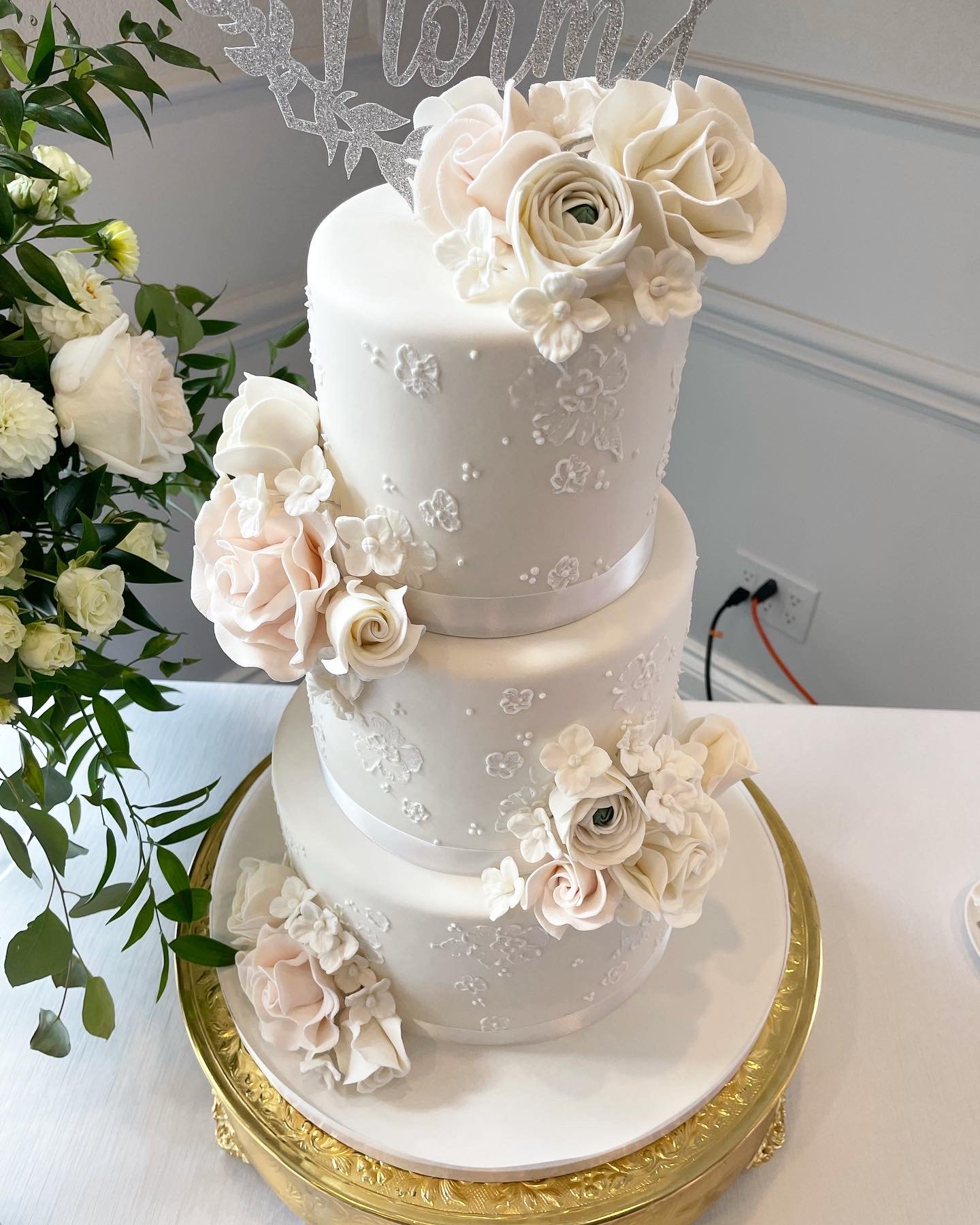 A three-tiered white wedding cake decorated with white flowers and floral patterns, topped with a cluster of white roses.
