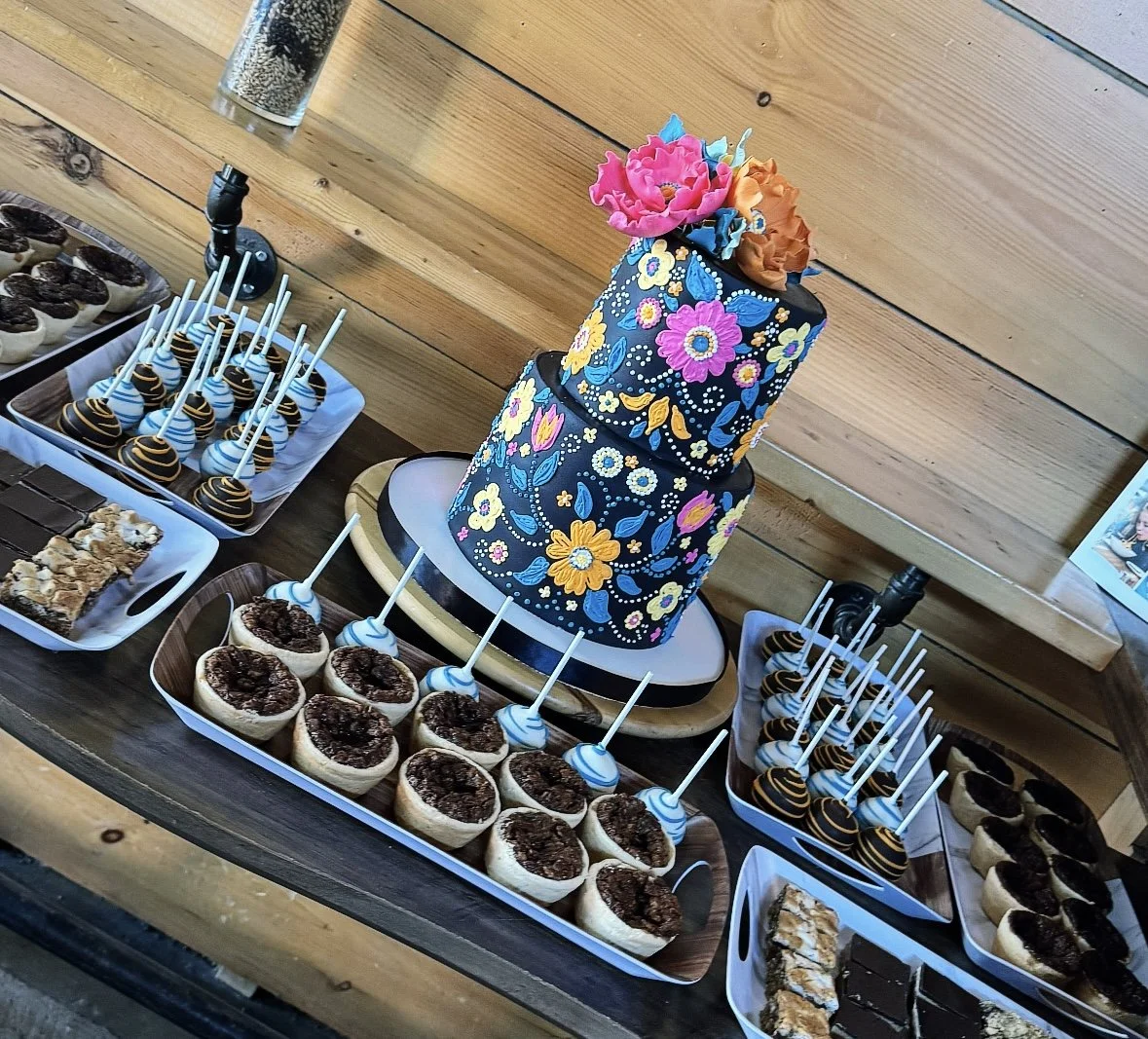 A dessert table featuring a two-tiered cake with floral decorations, surrounded by trays of mini cheesecakes with cherry topping and cake pops. The background has a wooden wall.