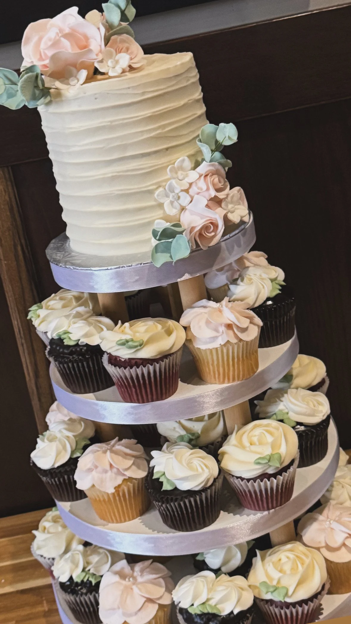 A tiered display of cupcakes with cream frosting and floral decorations, topped by a large white frosted cake with pastel flower decorations.