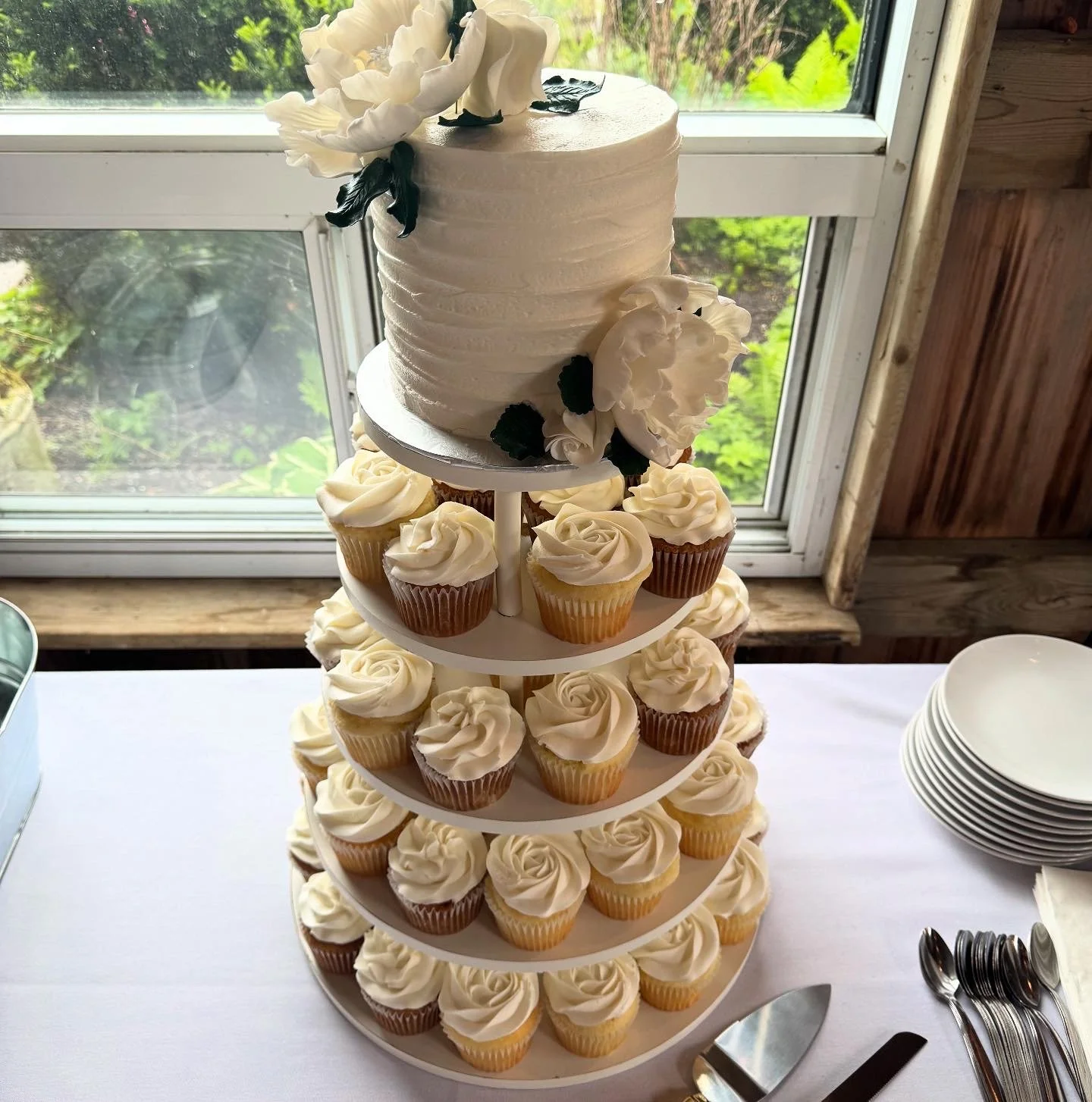 A tiered cupcake stand with a white cake on top and vanilla cupcakes with white frosting underneath.