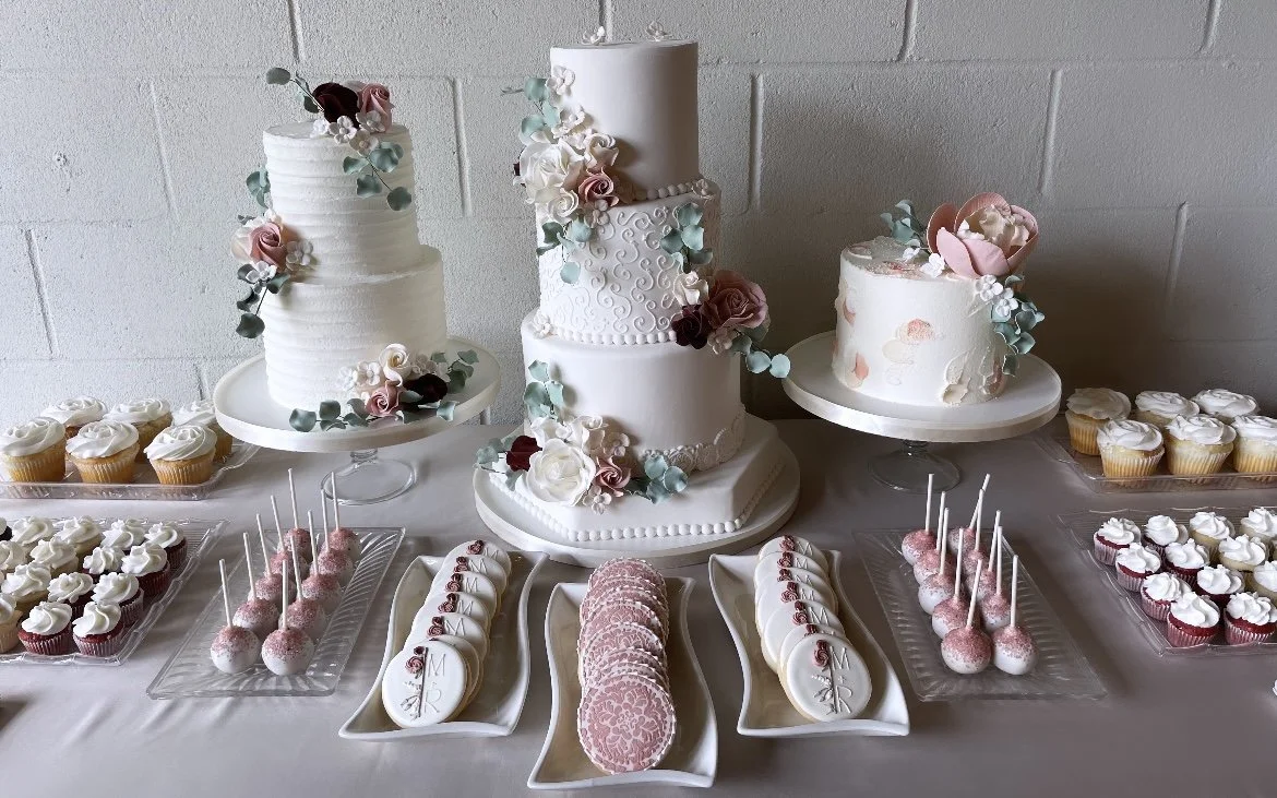 Assorted decorated cakes and cupcakes on a table, including a three-tier wedding cake with floral decorations, a single-tier cake, and various cupcakes with white frosting, pink, and red details, and cake pops, for a celebration.