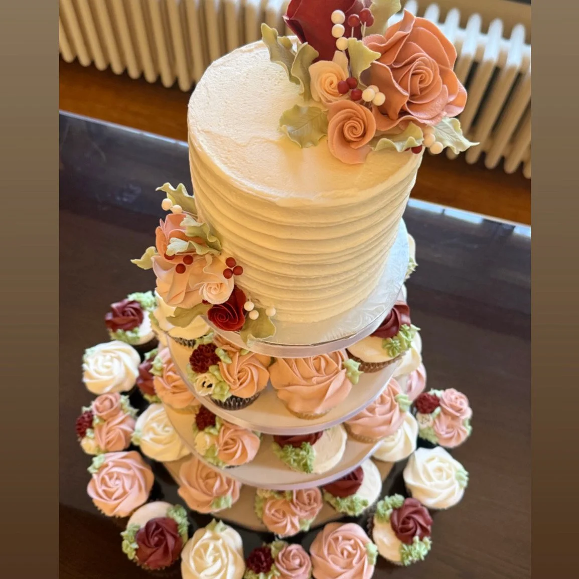 A tall, elegant wedding cake decorated with pink and white roses, green leaves, and small red berries, surrounded by matching cupcakes with rose designs, arranged on a multi-tiered stand.