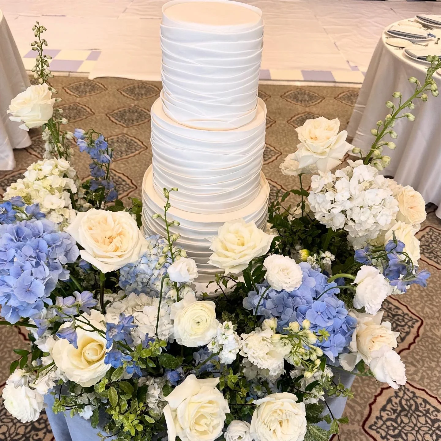 A tall, multi-tiered white wedding cake with a textured, horizontally striped design, surrounded by white and light blue flowers, including roses and hydrangeas, on a decorated table at a wedding reception.