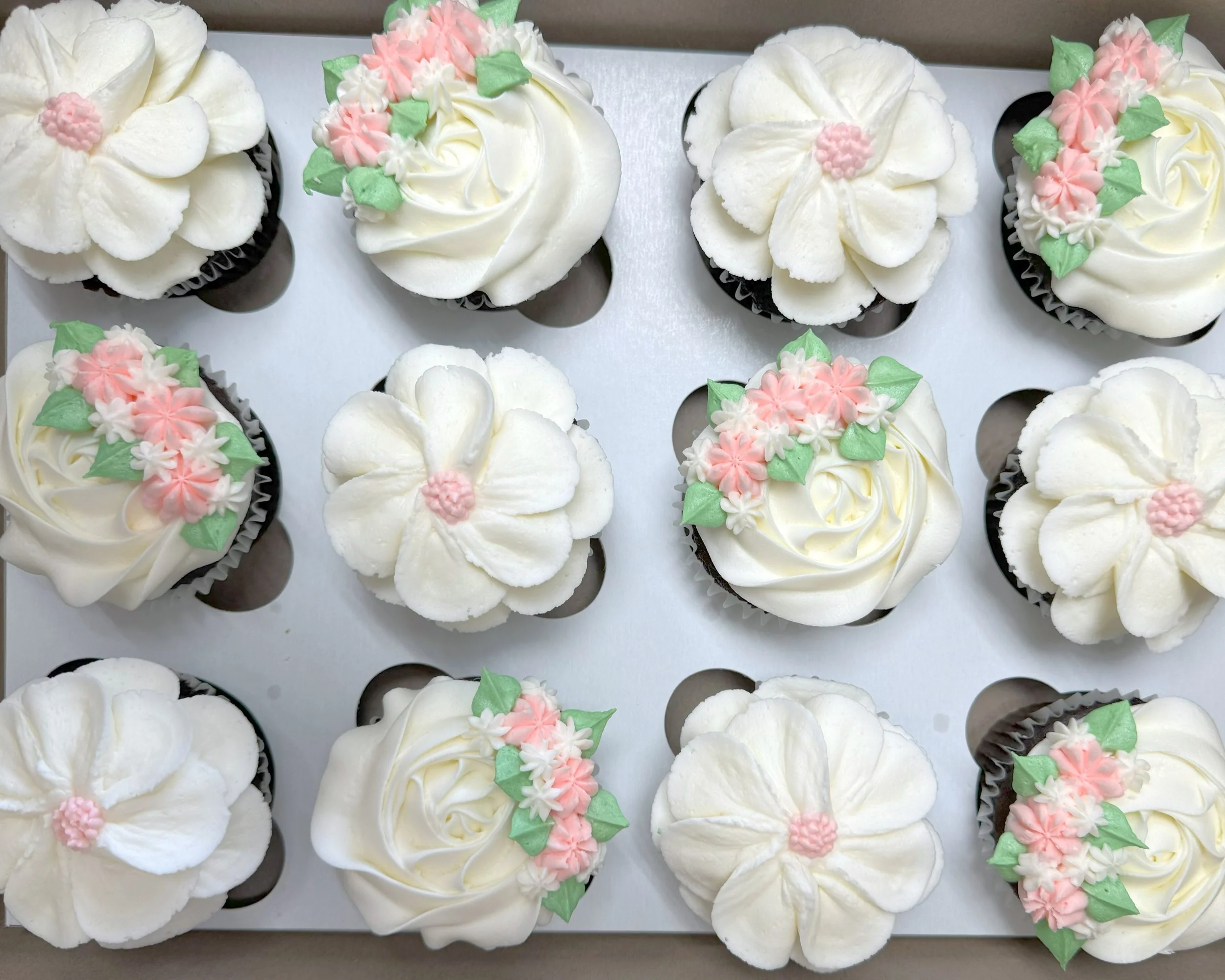 Cupcakes decorated with white frosting, pink and white floral icing, and green leaves.