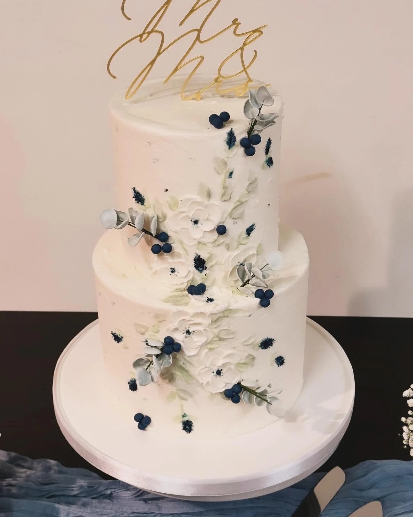 White, two-tiered wedding cake decorated with white and dark blue flowers, berries, and gray leaves.
