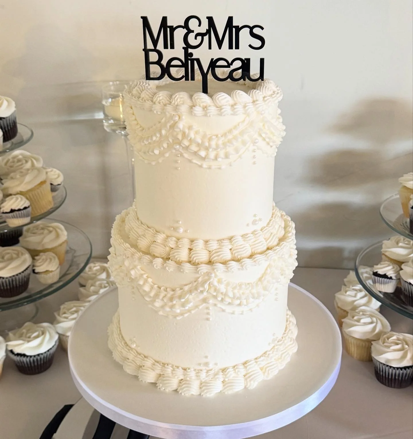 A three-tier white wedding cake decorated with piped icing and pearl-like accents, topped with a black cake topper reading 'Mr & Mrs Beliveau,' surrounded by cupcakes on glass stands.