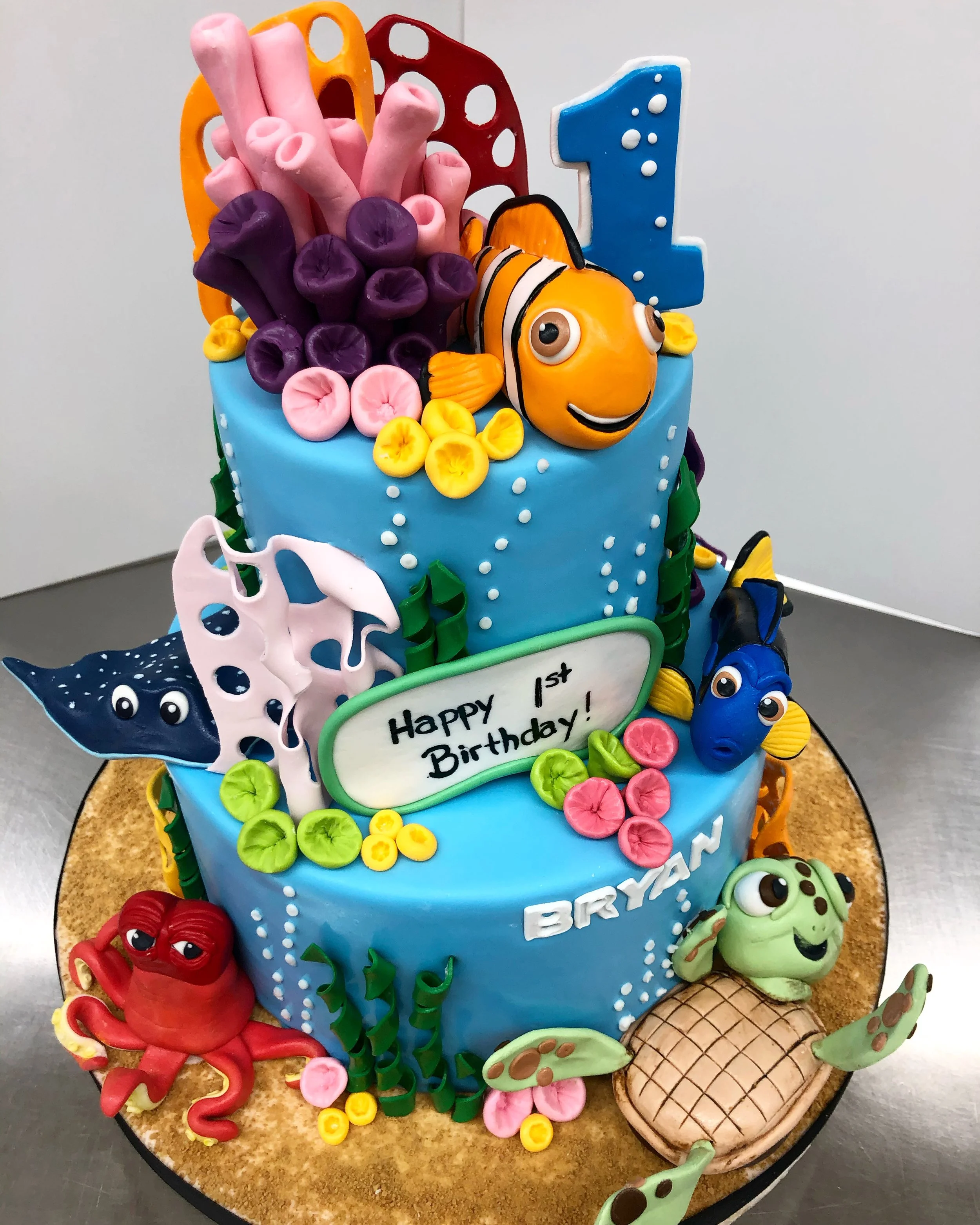 Two-tiered cake decorated with aquatic theme, featuring fondant sea animals like fish, octopus, turtle, and coral.