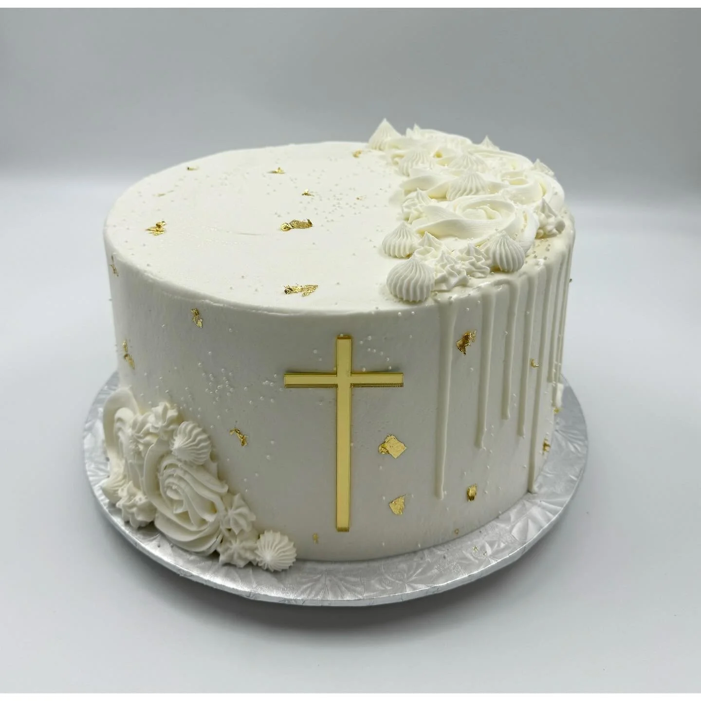 White religious-themed cake decorated with gold cross and gold accents, piped white frosting, and gold leaf embellishments on a silver cake board.