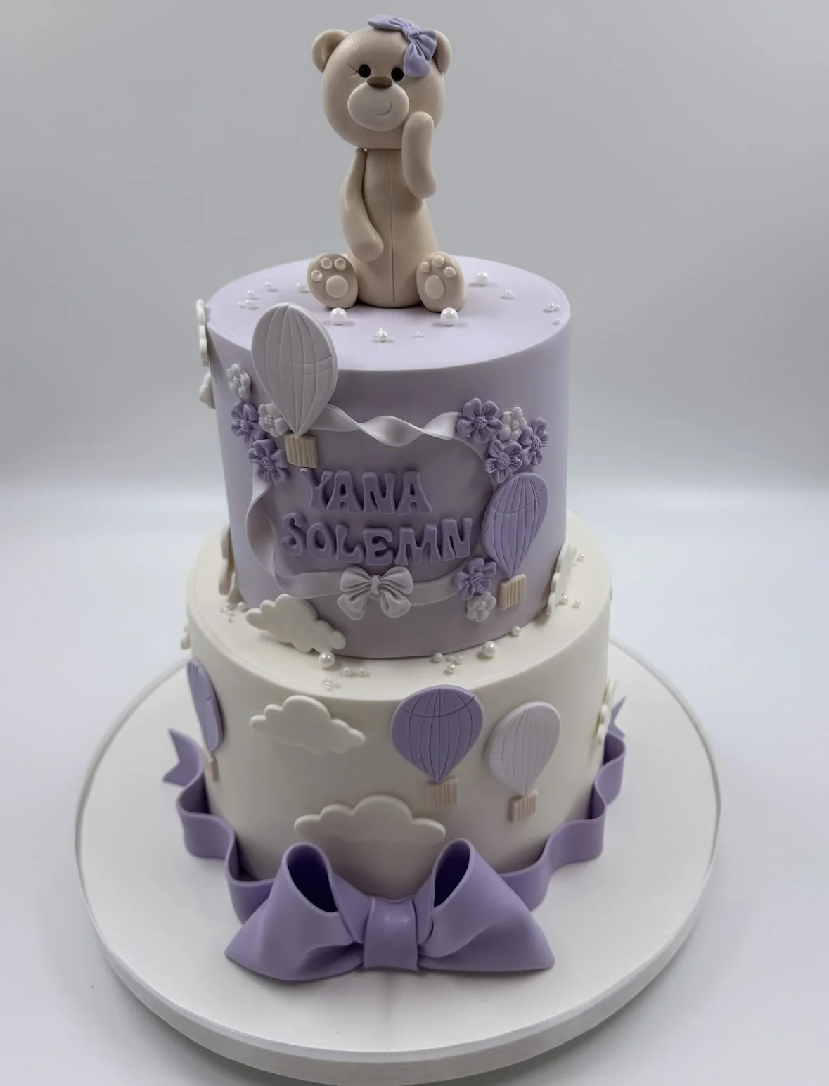 Three-tiered birthday cake with purple and white decorations, topped with a teddy bear figurine wearing a purple bow.