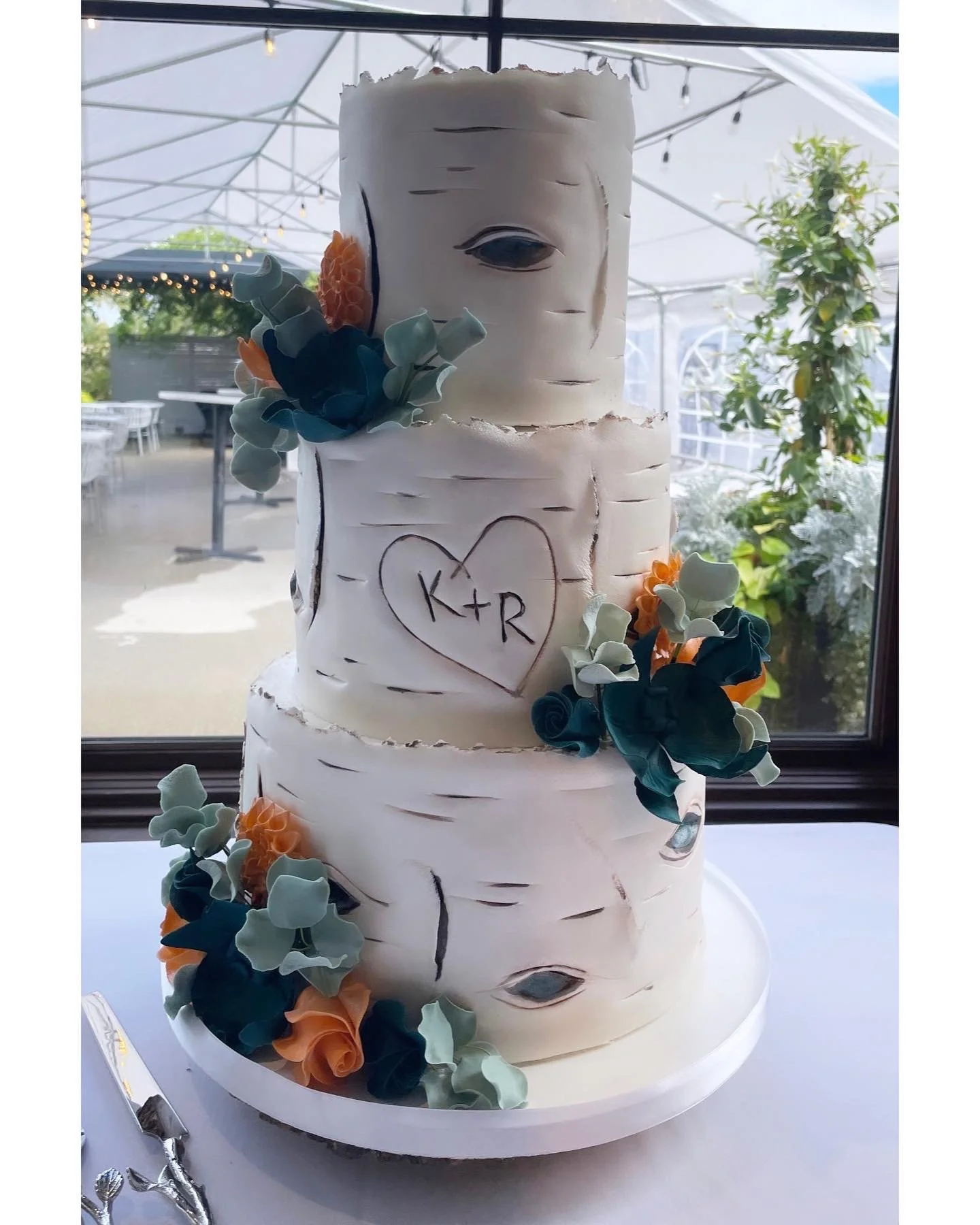 Three-tier wedding cake designed to look like a birch tree, decorated with fondant flowers and a heart with initials 'K+R', featuring a face with a single eye on the top tier, set in a bright, outdoor reception area.