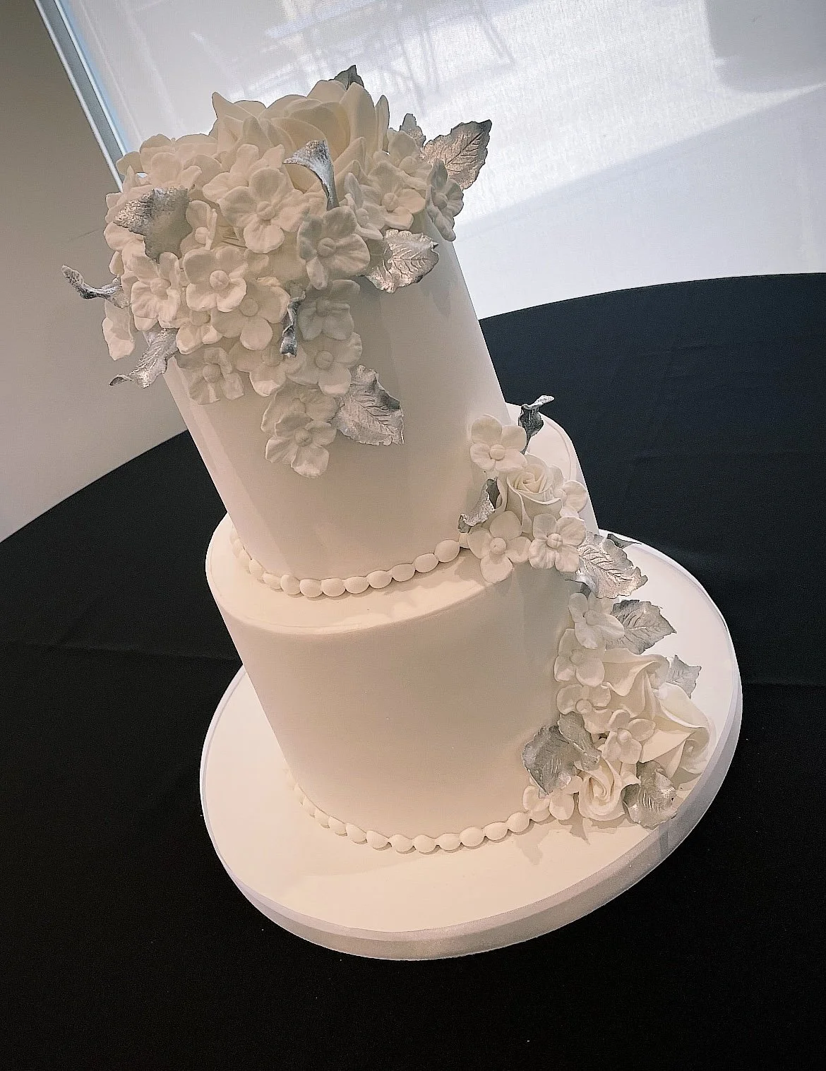A two-tier white wedding cake adorned with white flowers and silver leaves, placed on a white cake board.