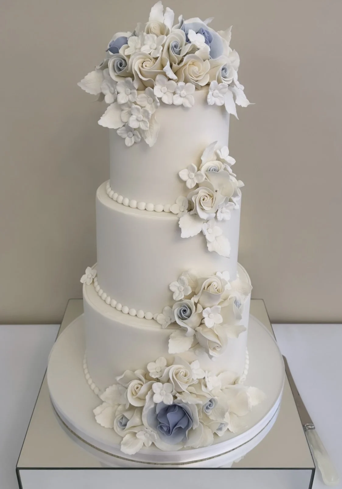 Elegant four-tier white wedding cake decorated with white and light blue sugar roses and small white flowers, with a cascading floral arrangement on top and around the tiers.