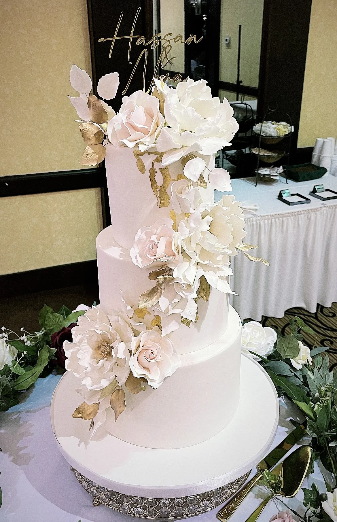 A three-tiered white wedding cake decorated with large white and light pink flowers, including roses and peonies.