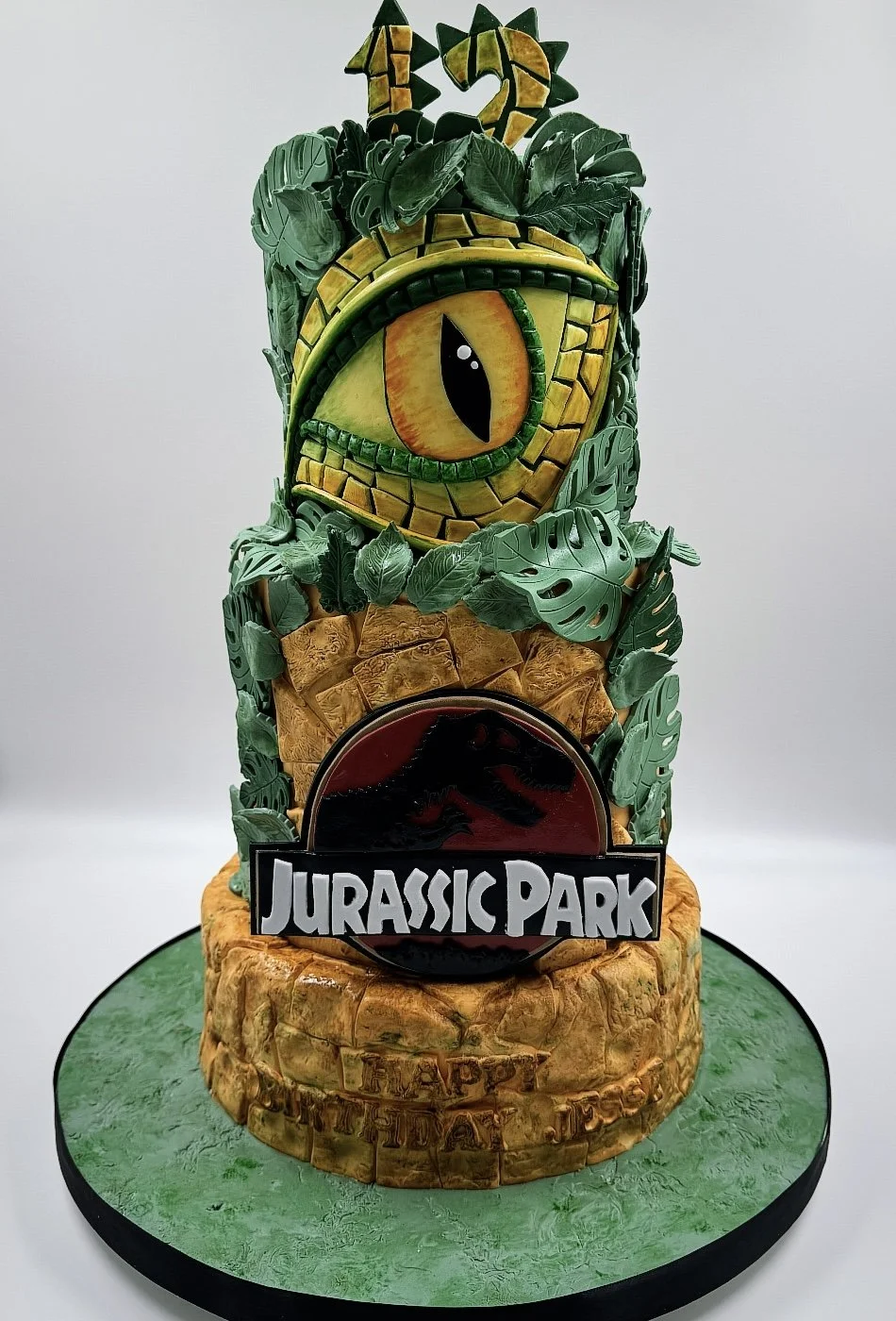 A Jurassic Park themed cake featuring a large eye of a dinosaur, surrounded by green leaves, with the Jurassic Park logo in front, on a green base.