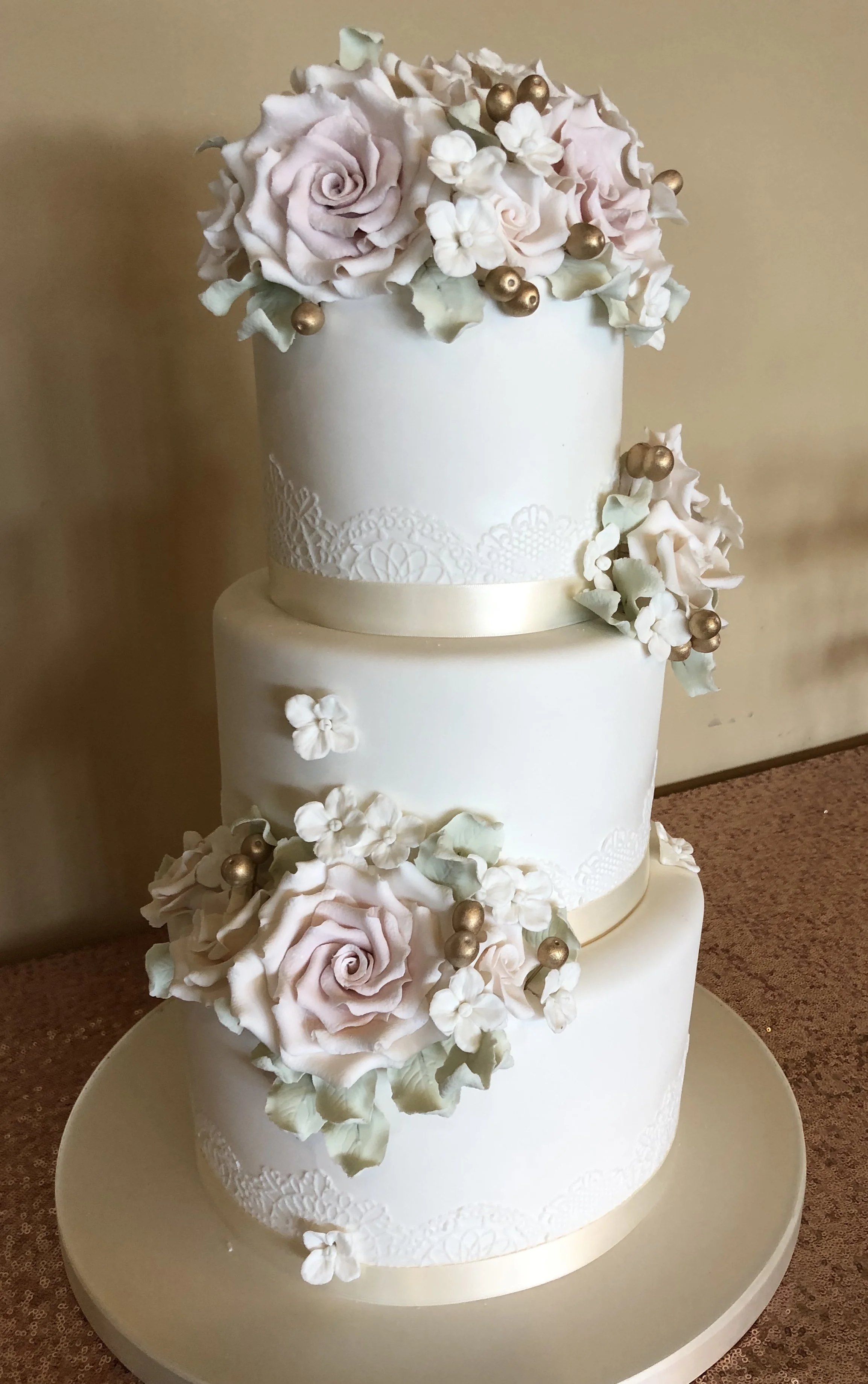 Three-tier white wedding cake adorned with pink roses, white hydrangeas, gold spherical accents, and ribbon decorations.