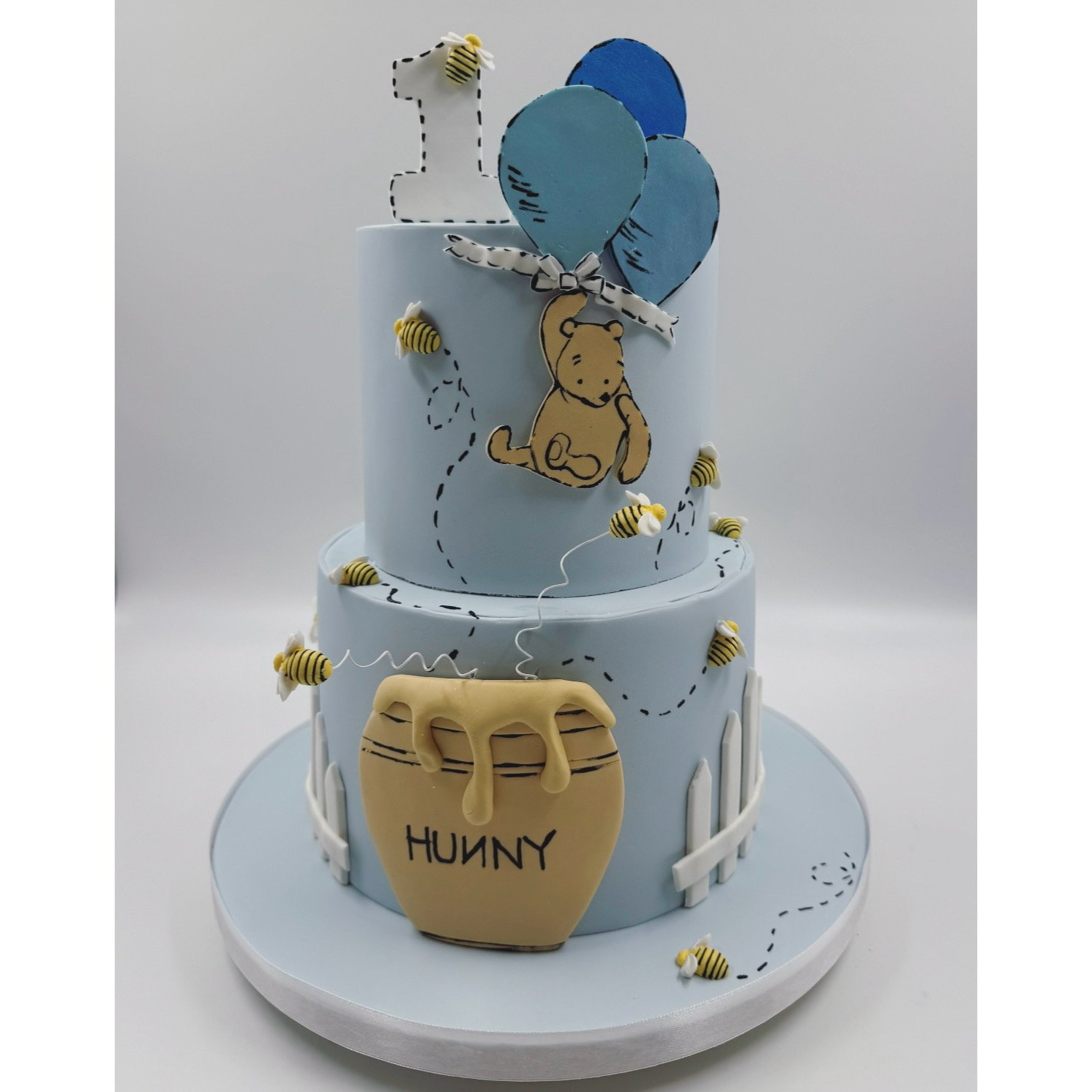 Two-tiered light blue birthday cake with bear-themed decorations, including a puppy drawing, bees, a honey pot labeled 'HUNNY', and a number one candle with a bee on top. The cake also has white picket fences and balloon decorations.