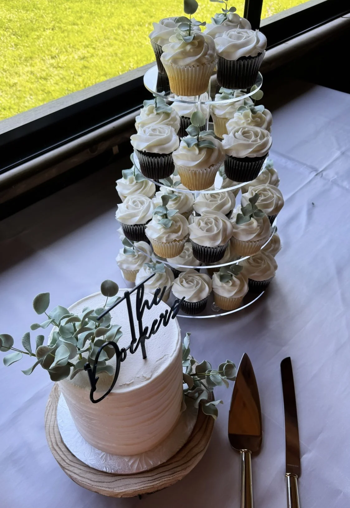 A tiered display of cupcakes with white frosting and small green leaves, a round white cake with greenery.