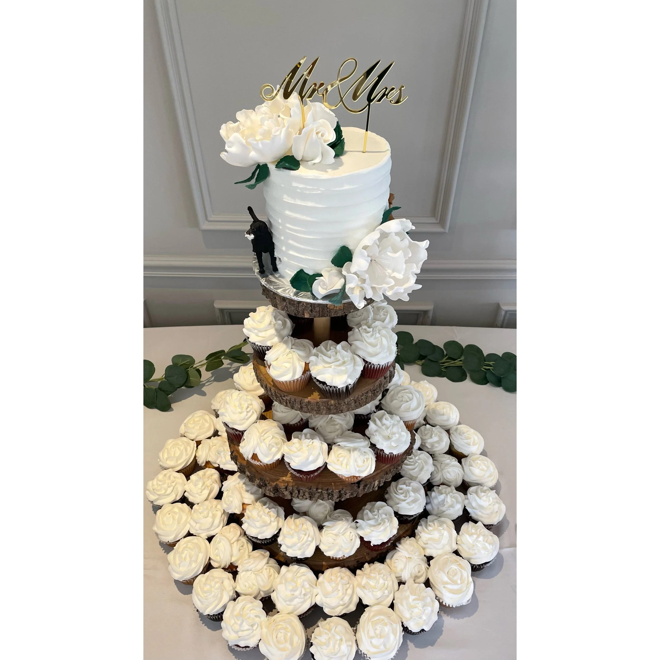 Wedding cake decorated with white flowers and surrounded by cupcakes on a tiered wooden stand with additional cupcakes on a table.