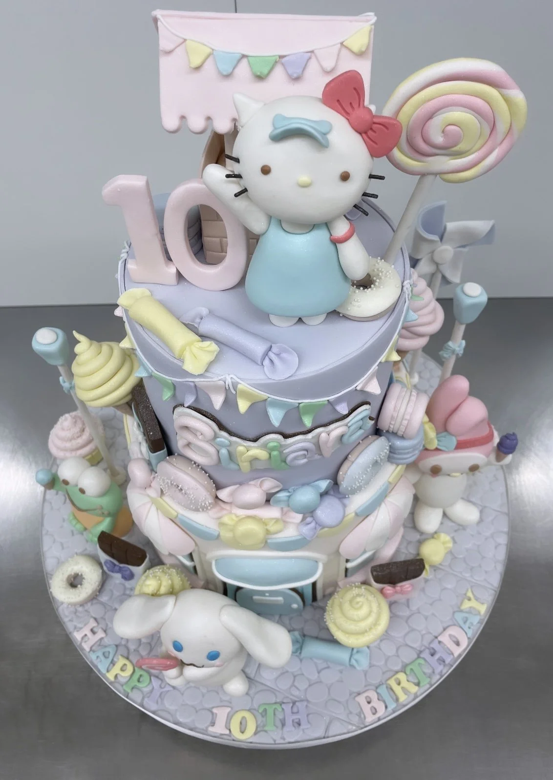 A pastel-colored birthday cake decorated with Hello Kitty figures, candies, featuring a main Hello Kitty figure on top holding a lollipop and waving.