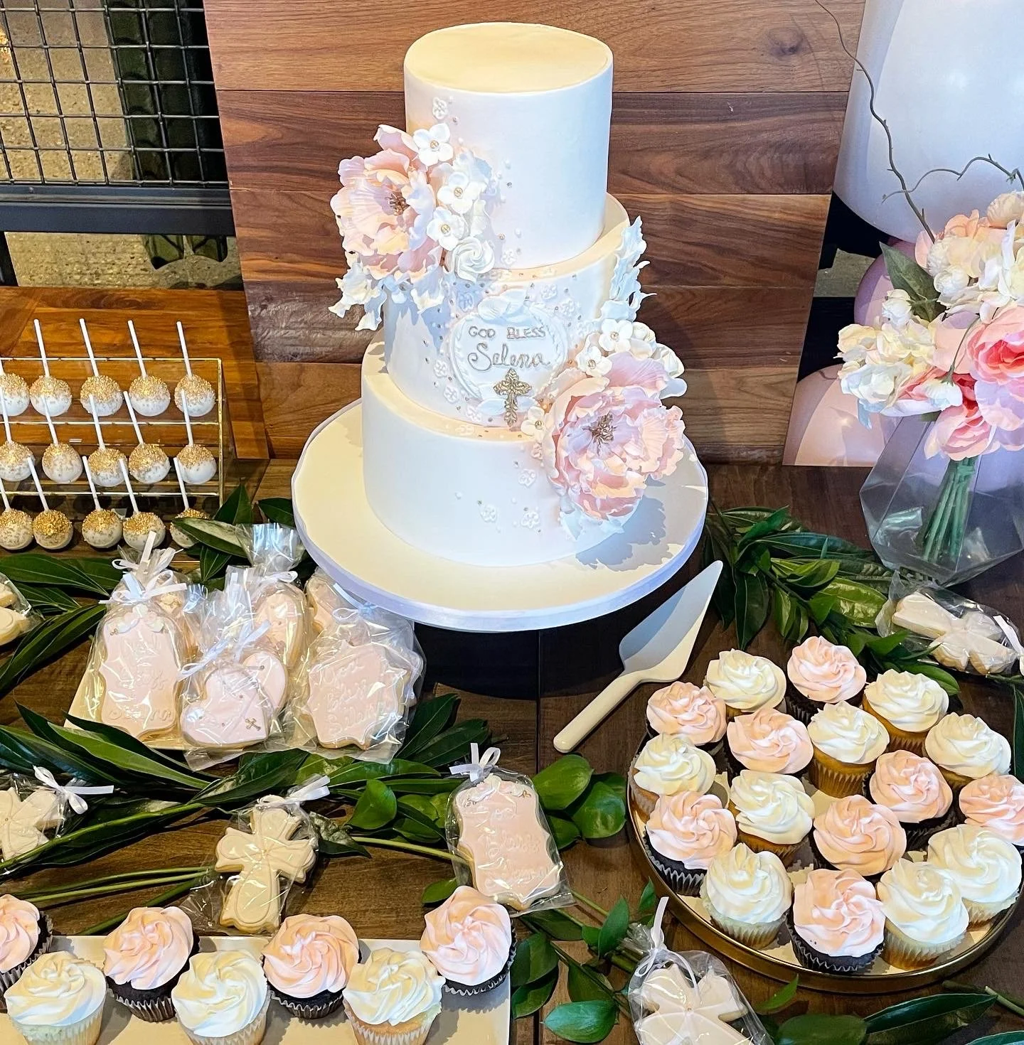 A three-tier white wedding cake decorated with pink and white flowers, placed on a white cake stand. The cake has a small plaque labeled "God Bless Solana" with a cross. Surrounding the cake are cupcakes with pink and white swirled frosting, cookies 
