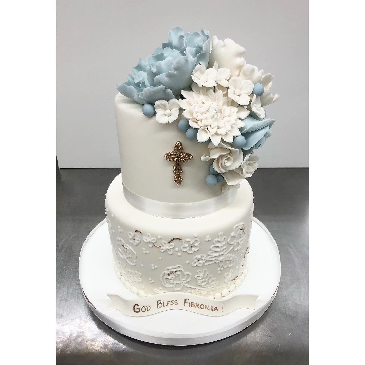Two-tiered white cake with floral decorations on top. The top tier features a gold crucifix attached to the white surface.