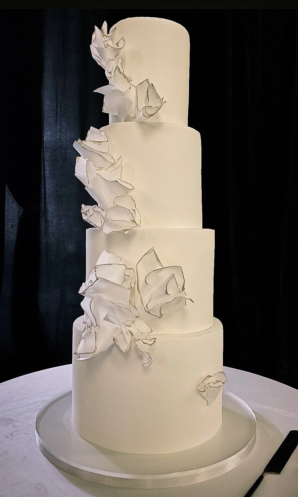 White four-tier wedding cake with crinkled white decorative flowers on each tier.