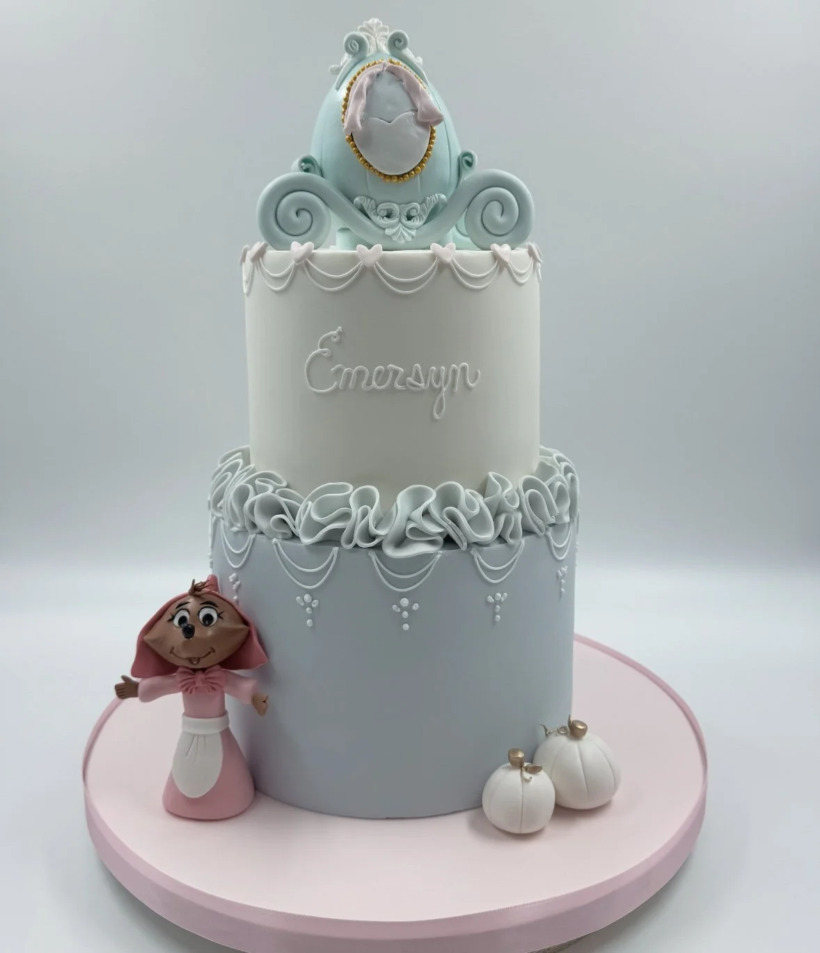 Two-tiered pastel-colored birthday cake featuring a character from 'Encanto' at the bottom and a decorative toy at the top, with pumpkins around the base, on a pink round cake board.
