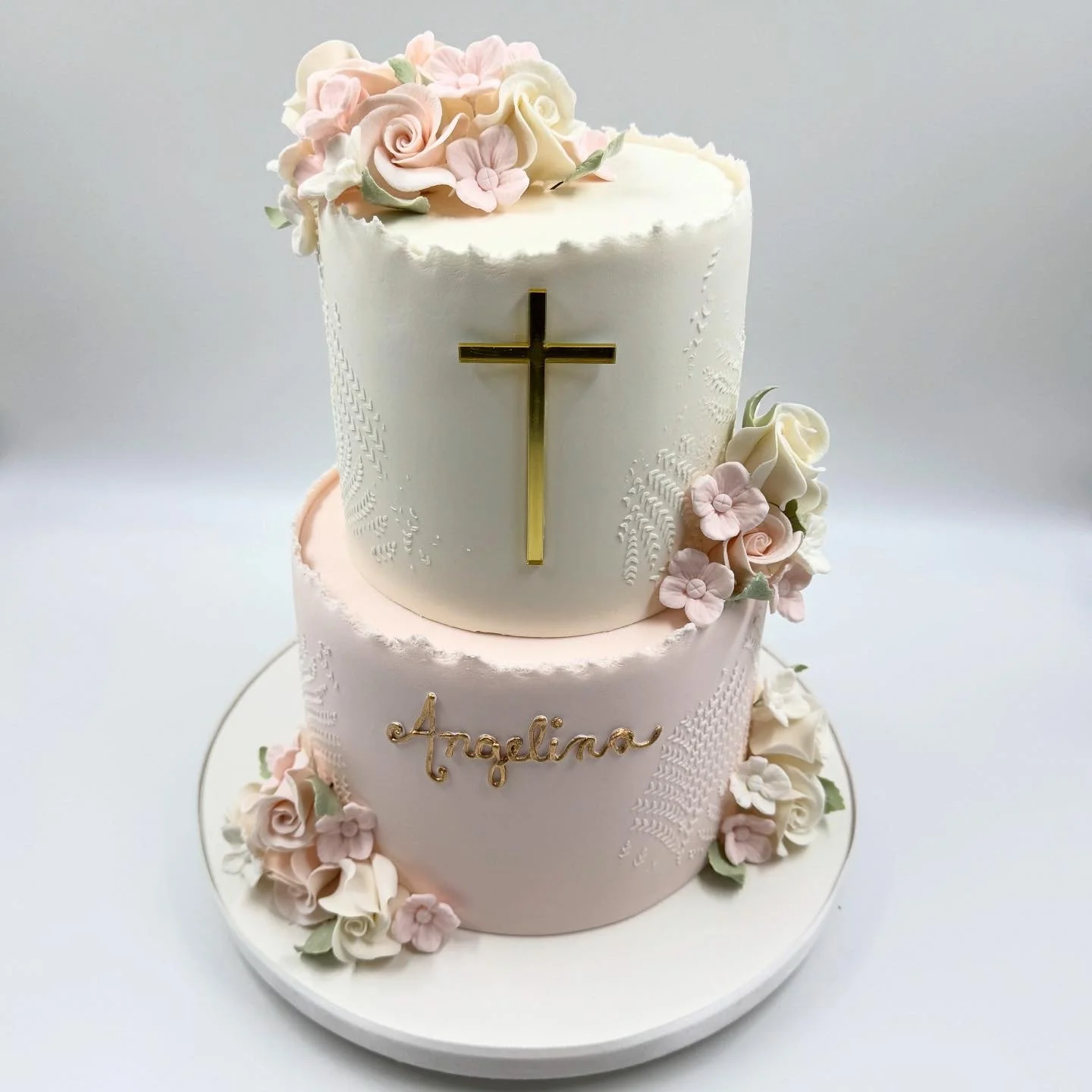 Two-tier white and pink wedding cake decorated with pink and white flowers, a gold cross on the top tier, and the name 'Angelina' in gold on the front of the bottom tier, displayed on a white stand.