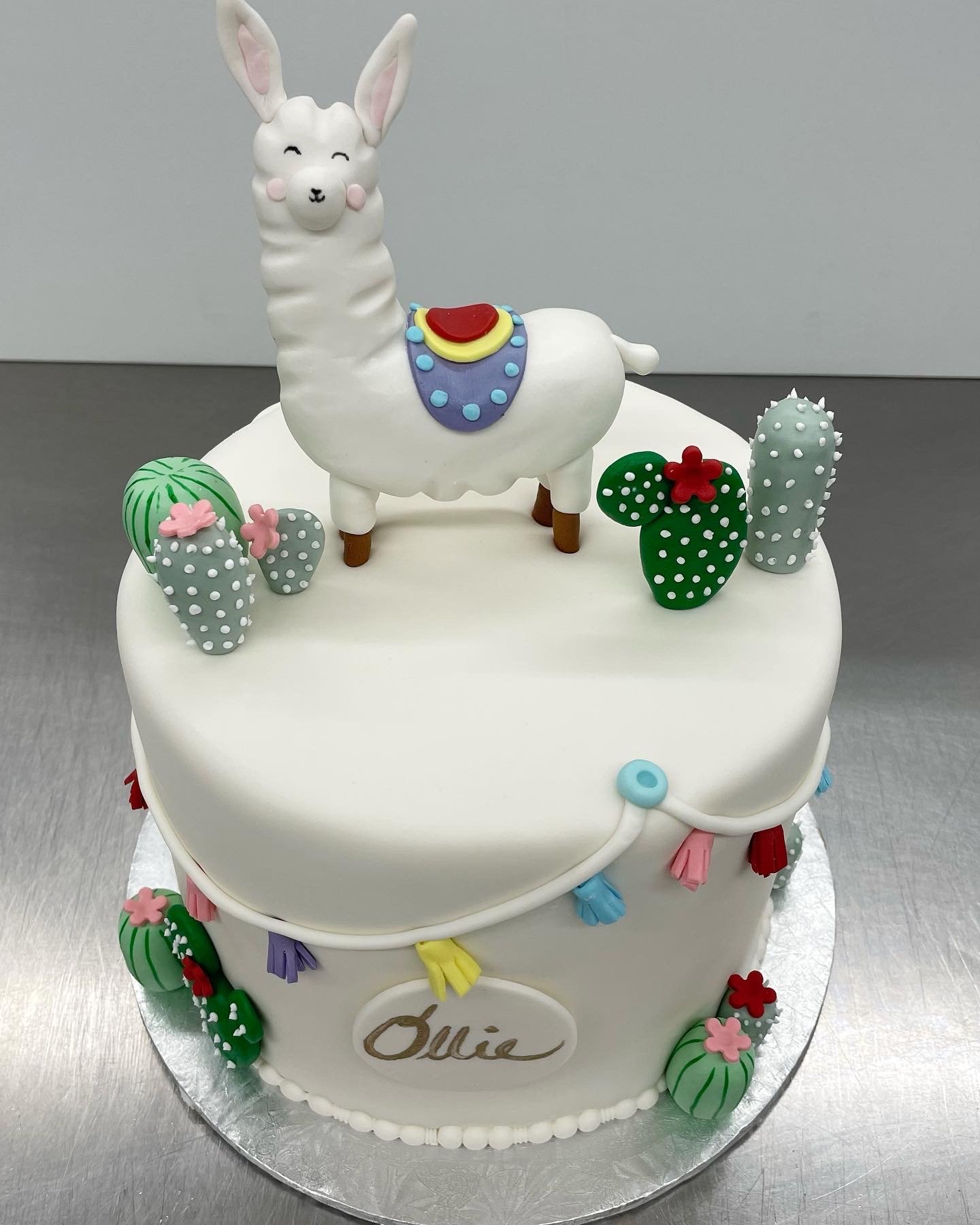A white birthday cake decorated with a llama figurine on top and various cactus-shaped decorations around it. The cake has colorful tassels.