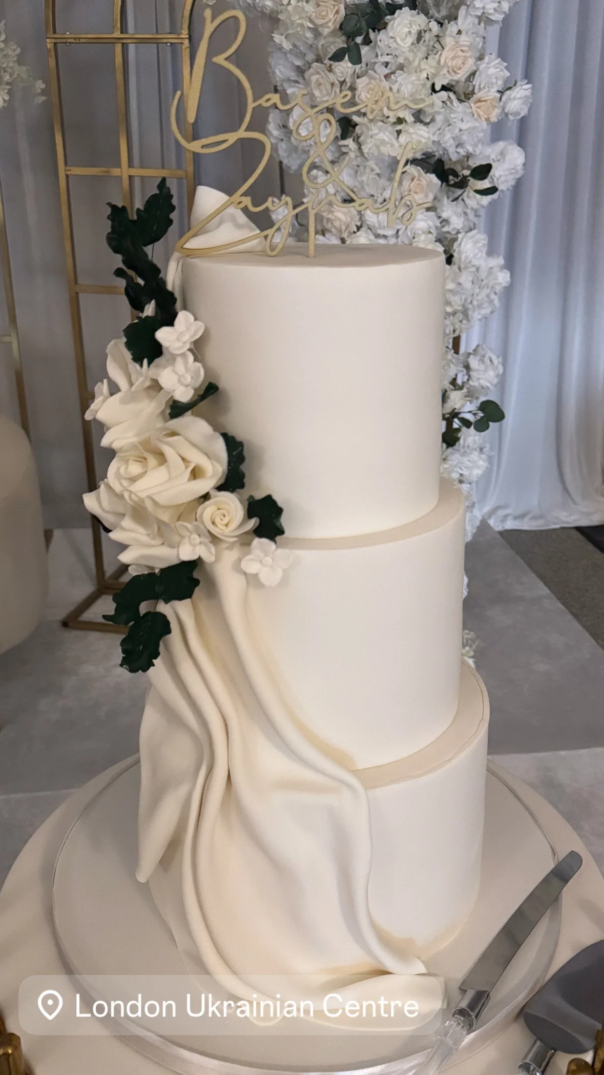 A three-tier white wedding cake decorated with a cascading arrangement of white flowers and dark green leaves, topped with a gold "Bride & Groom" cake topper, displayed at the London Ukrainian Centre.