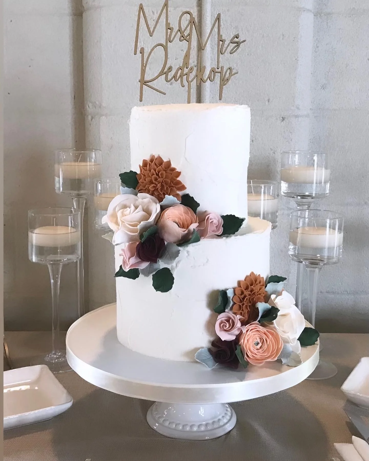 Two-tier white wedding cake with flower decorations, cake topper with the words "Mr & Mrs Padeh Kon" on top, surrounded by eight glass candle holders with white candles, on a round white cake stand.