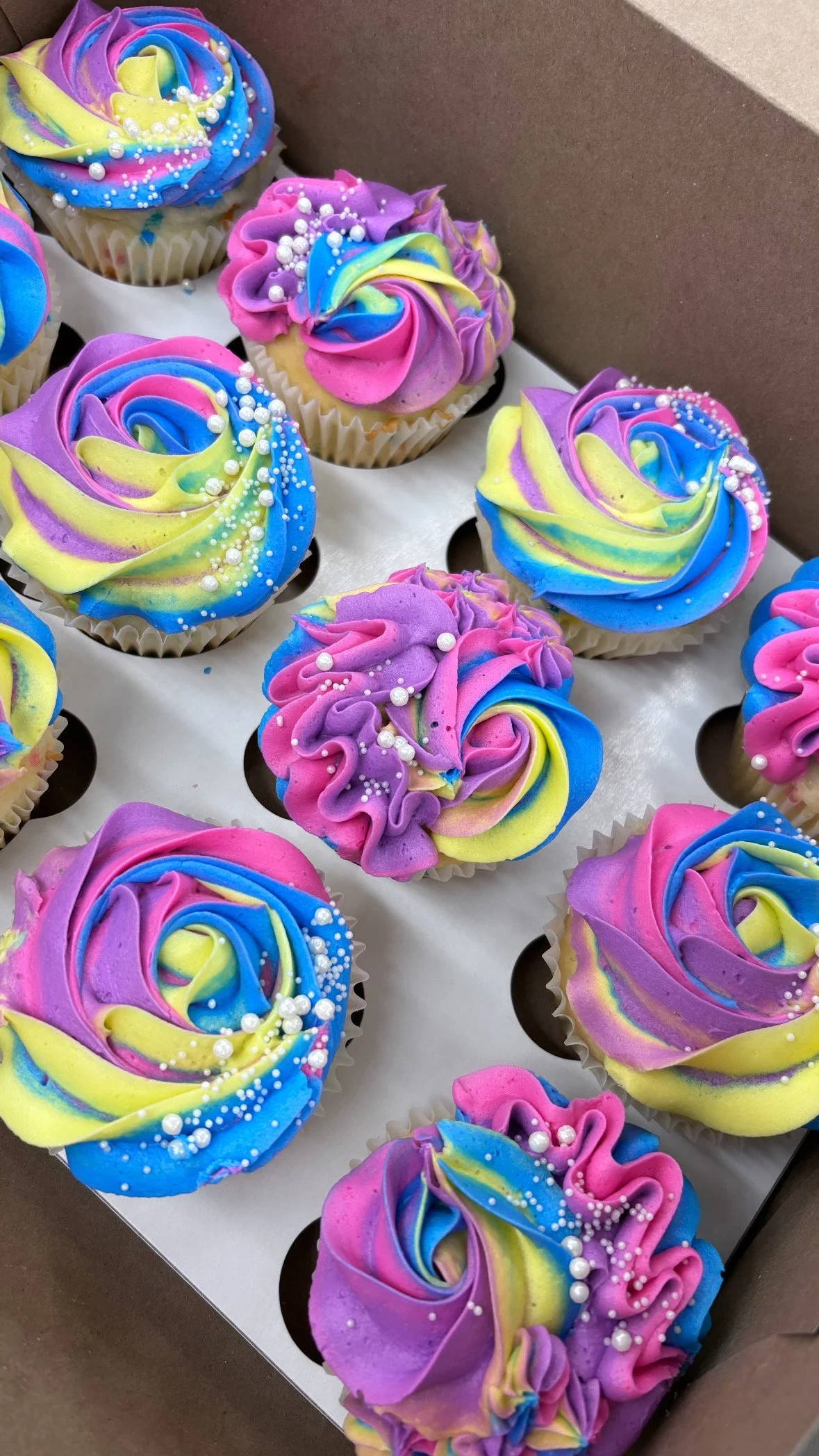 Colorful swirl frosting cupcakes in a box with pink, yellow, blue, purple, and green frosting, decorated with white pearl sprinkles.