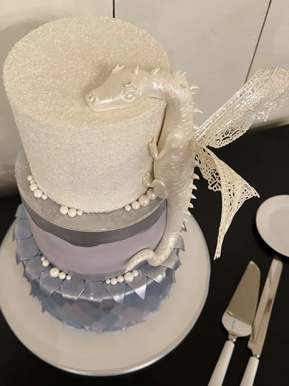 A three-tiered cake with a white, glittery, textured upper tier, a light purple middle tier, and a silver ruffled bottom tier. It features a white dragon with pearl-like beads on its body and silver lace wings, sitting on the side of the top tier.