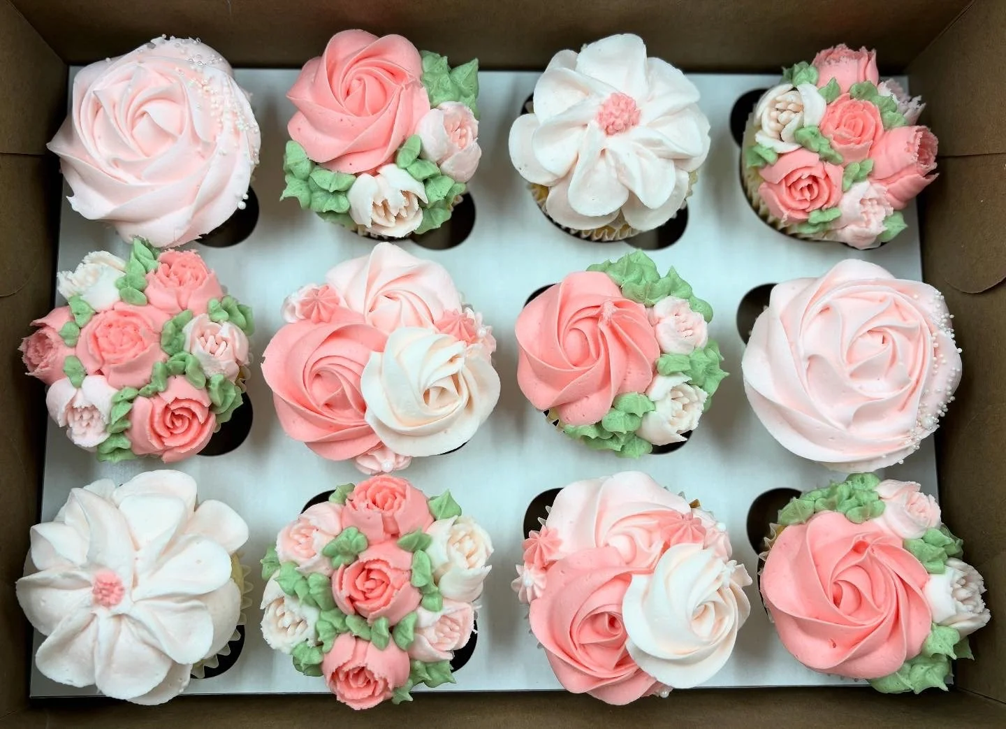 A box of twelve cupcakes decorated with pink and white buttercream roses and floral designs.