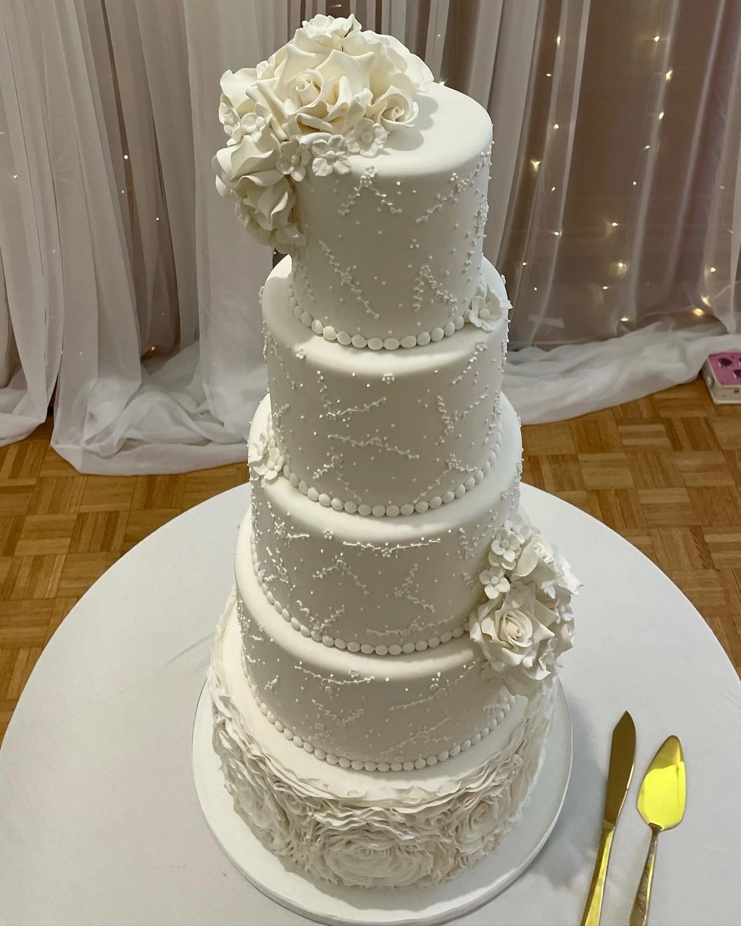 A tall, four-tier white wedding cake decorated with floral accents, white roses, and intricate piping details, placed on a white round table with a gold cake knife and server beside it.