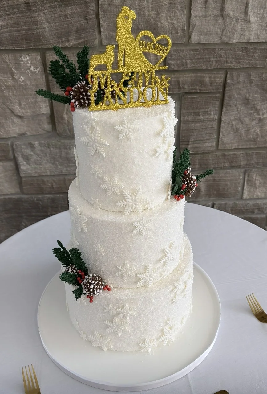 Three-tier white wedding cake decorated with snowflake patterns, holiday greenery, pinecones, and red berries, topped with a gold glitter cake topper reading "Love is Sweet" and "W & S London".