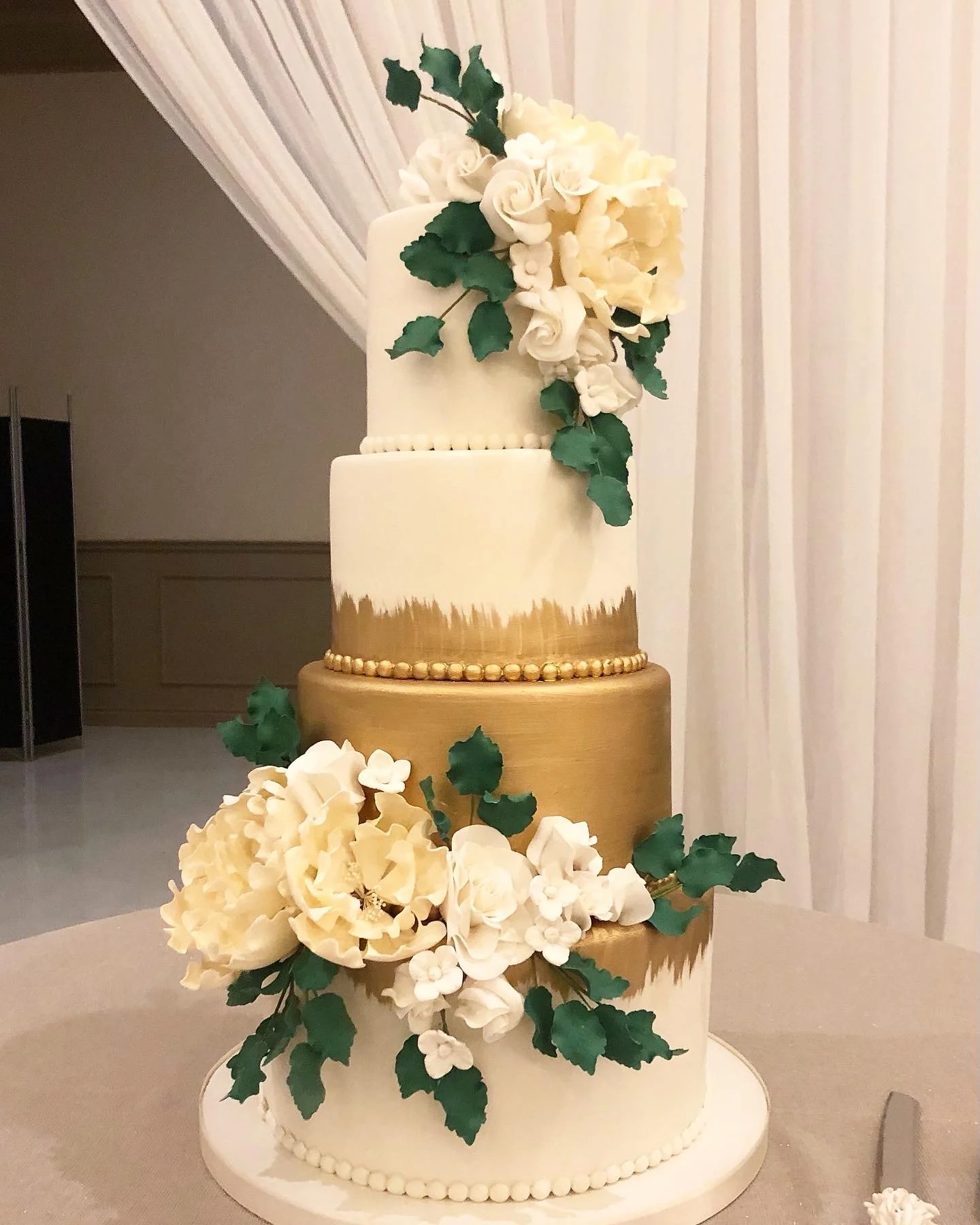 A three-tiered wedding cake decorated with white flowers and green leaves, featuring a gold and white color scheme, with floral arrangements on top.