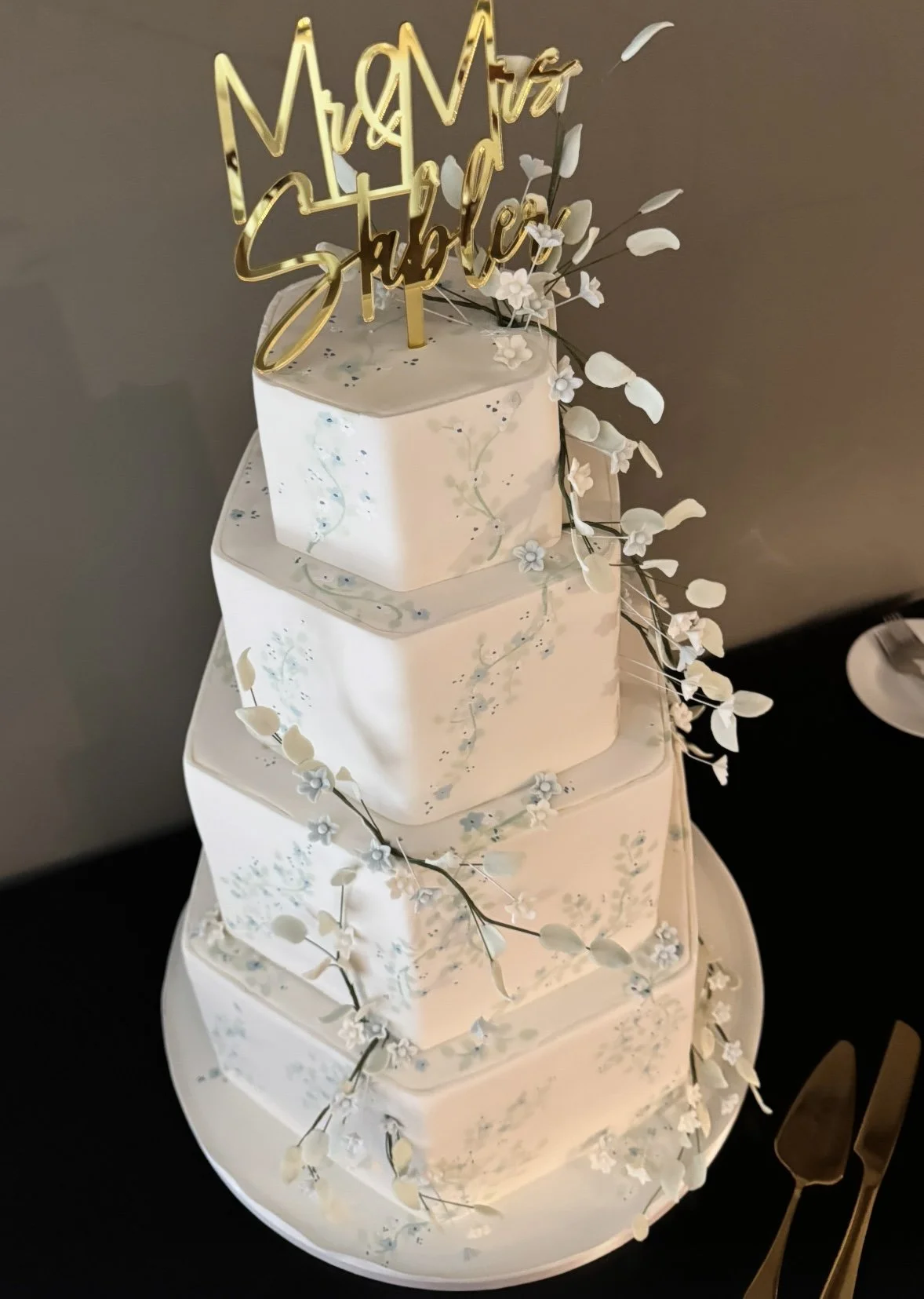 Four-tiered white wedding cake with delicate floral decorations and a gold "Mr. and Mrs. Steele" topper, topped with white flowers and green leaves.