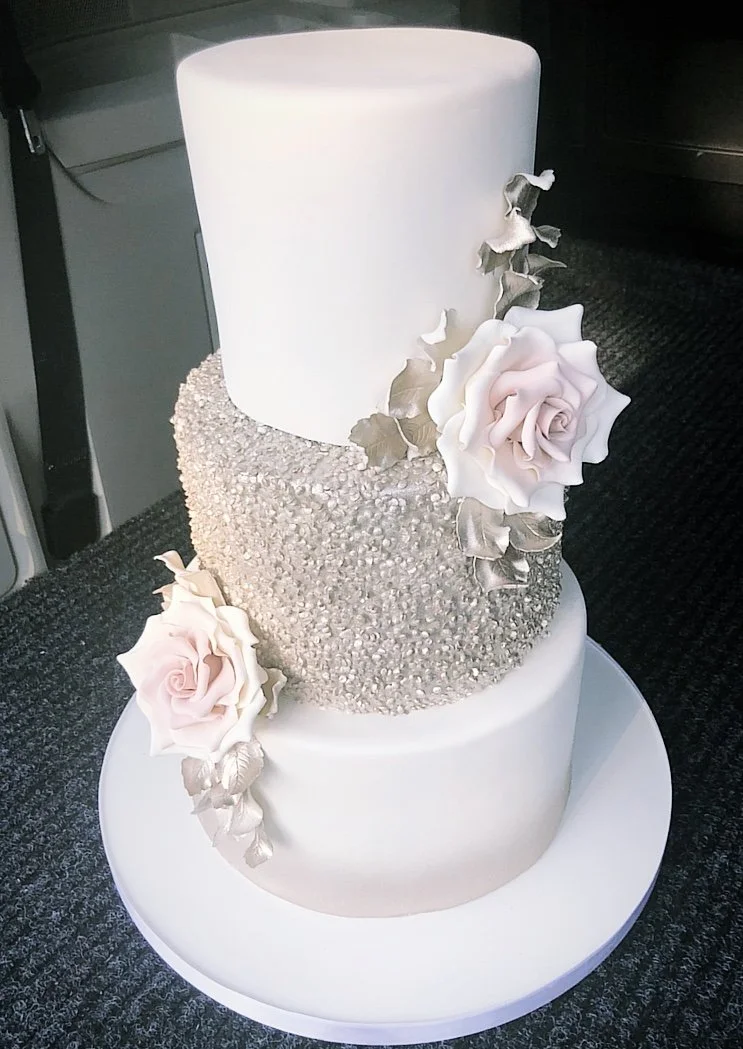 Three-tier wedding cake with white, textured, and smooth icing, decorated with pink and white roses and gold leaves.