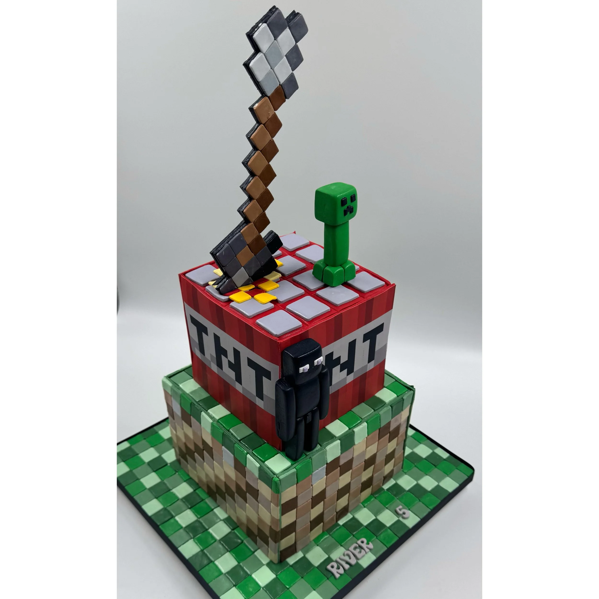 A decorated cake inspired by Minecraft featuring pixelated characters, a TNT block, a Creeper, a black Enderman, and a sword on top, with a Minecraft grass block and checkered base.