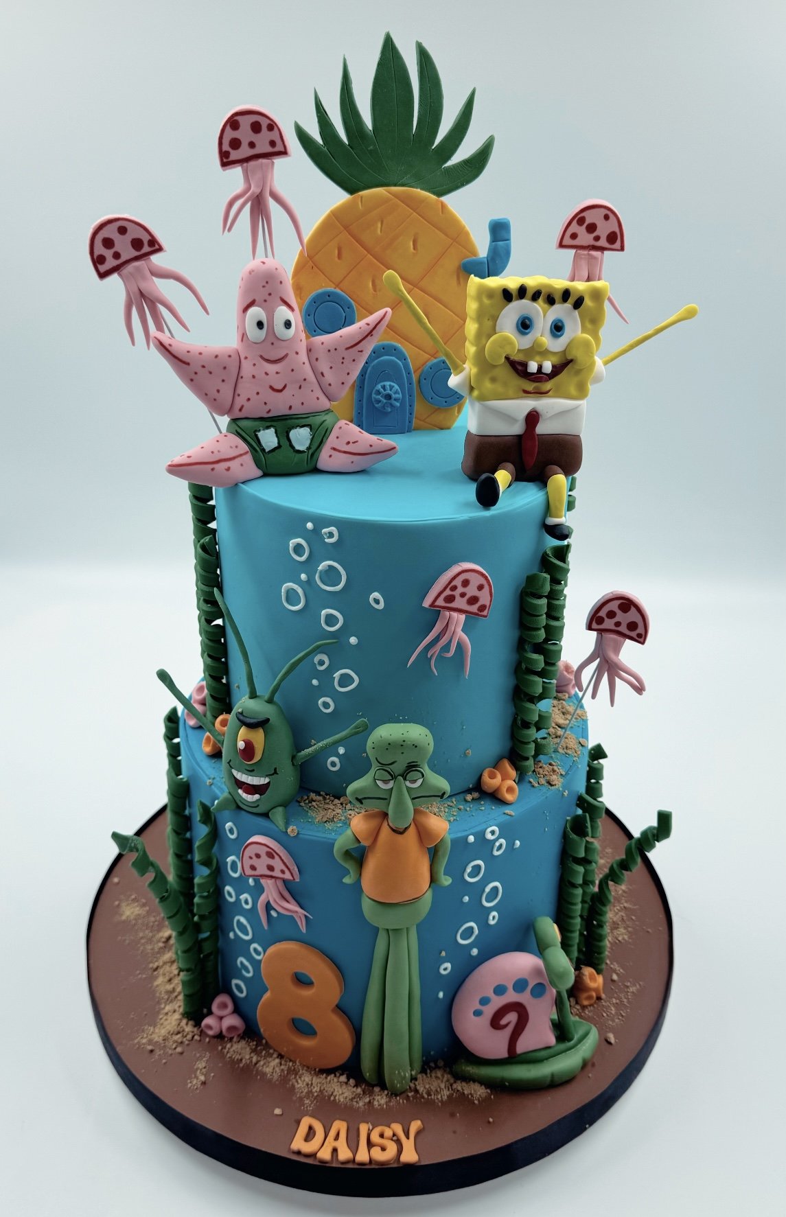 Colorful two-tiered birthday cake decorated with characters from SpongeBob SquarePants, including SpongeBob, Patrick, Squidward, and Plankton, with a pineapple top and sea-themed decorations.