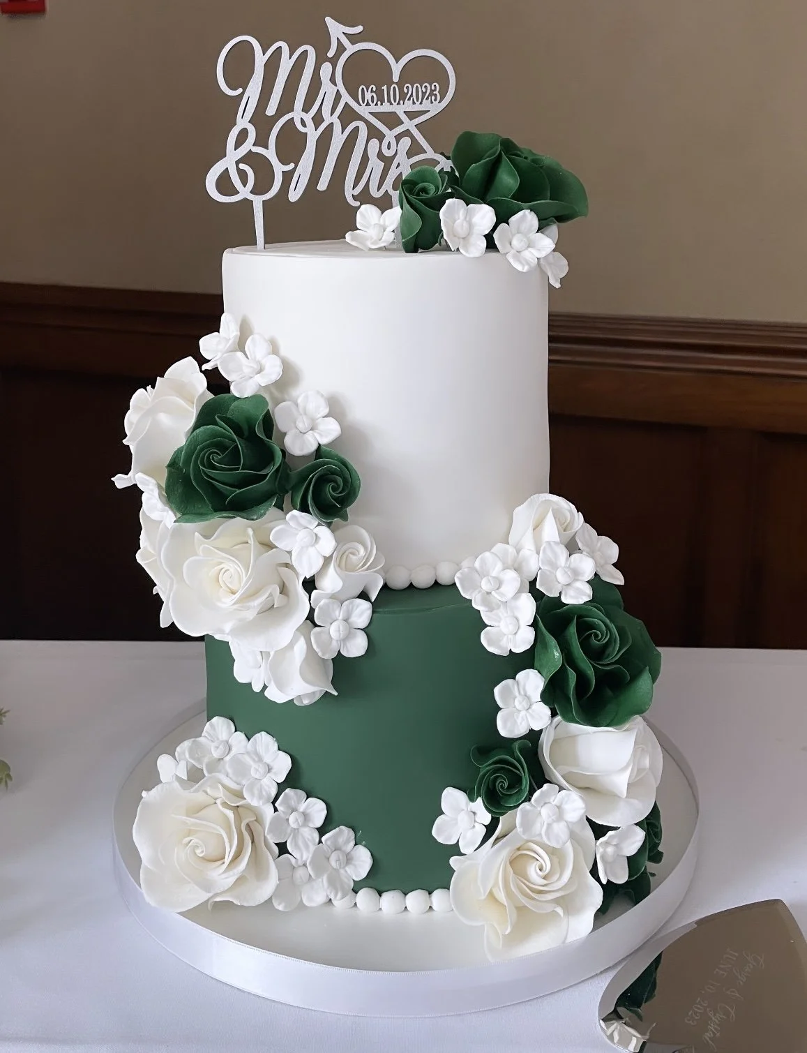 Two-tiered wedding cake with white and green fondant, decorated with white roses and small white flowers, topped with a cake topper reading "Mrs. & Mrs." and a date, 06.10.2023.