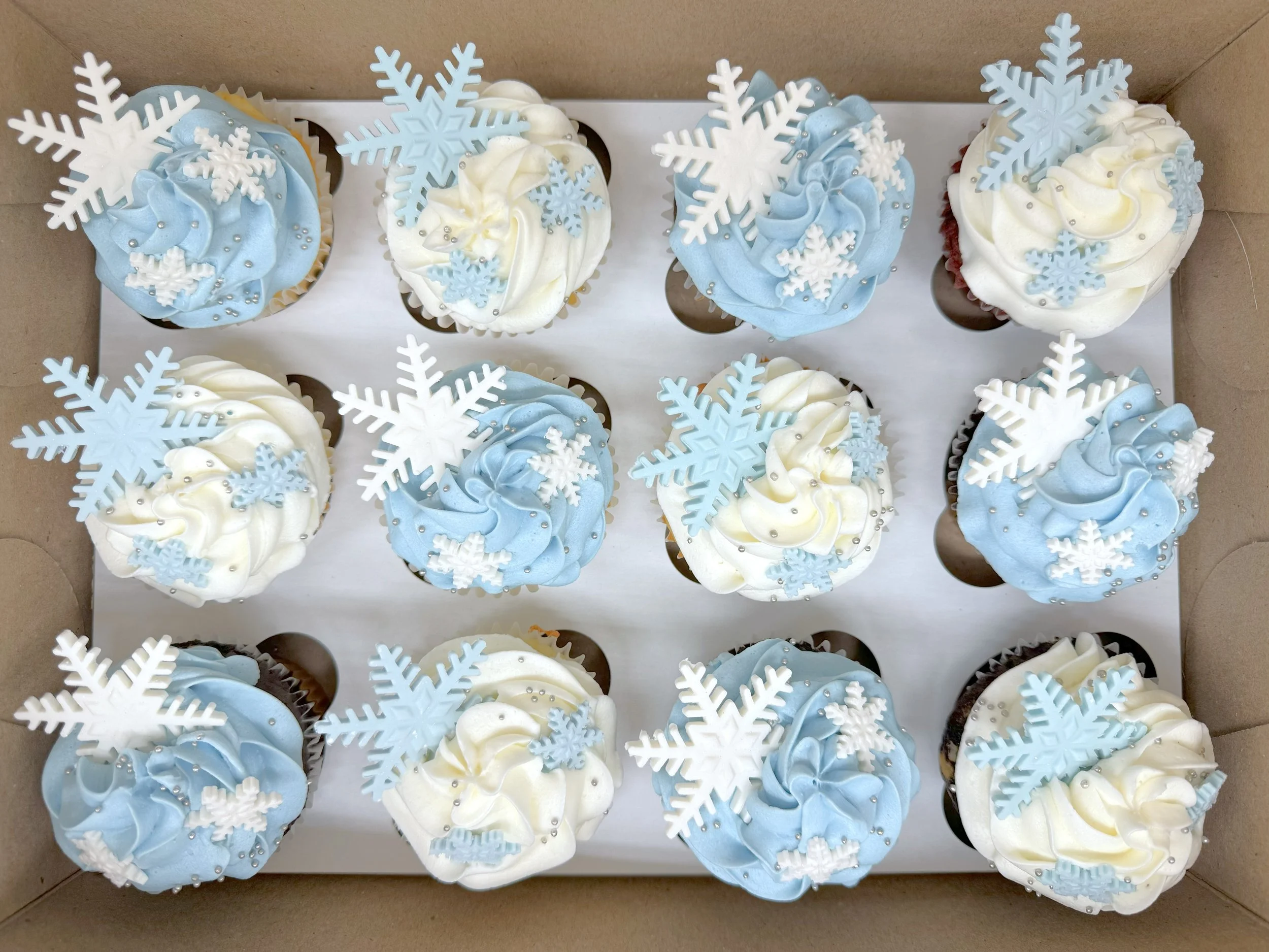 A box of twelve cupcakes decorated with white and blue frosting, snowflake toppers, and silver edible beads, arranged in a 3x4 grid.