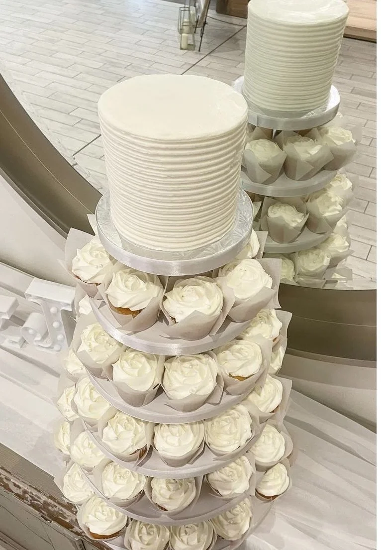 A large, stacked display of white cupcakes with white frosting, topped with two large white frosted cakes.