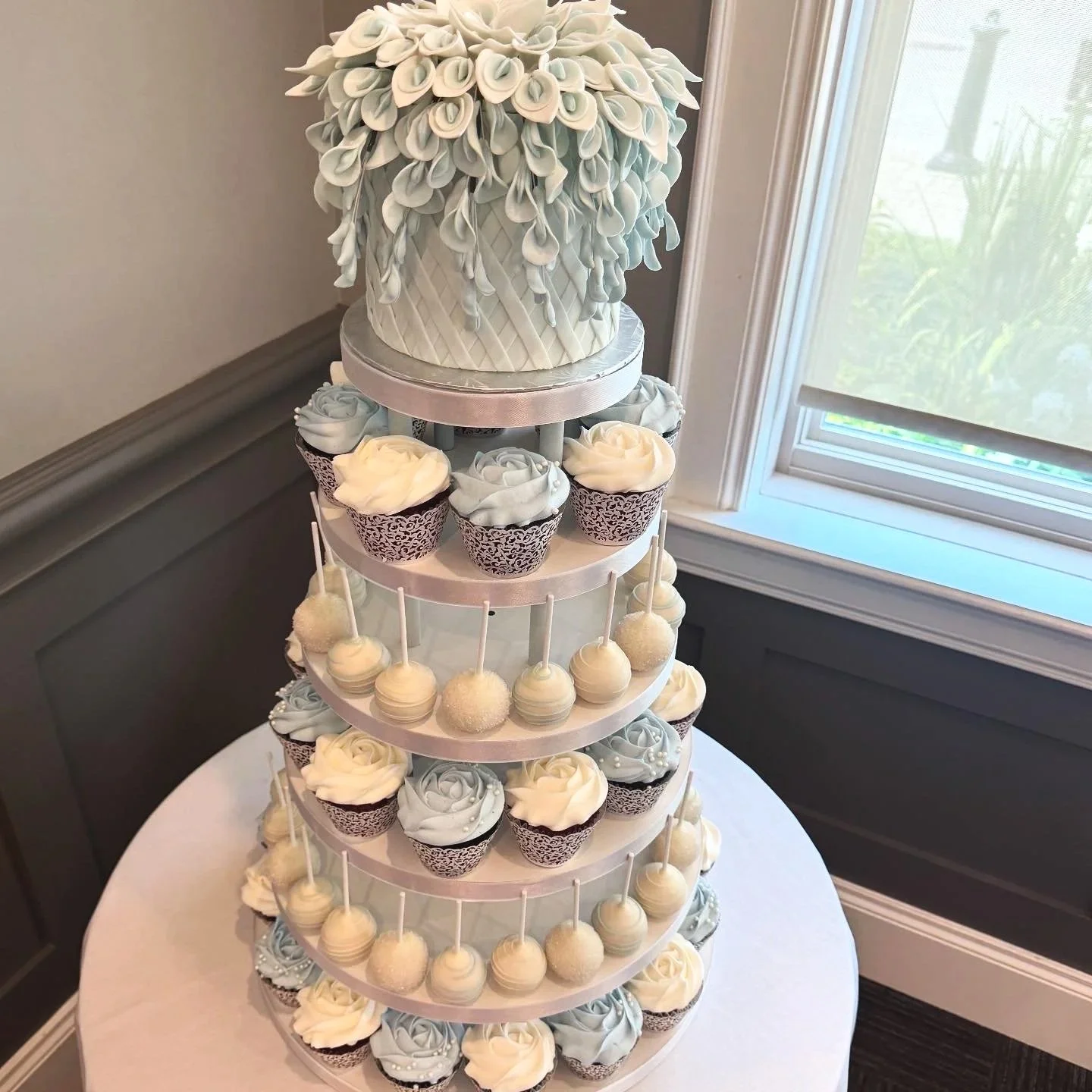 A multi-tiered dessert stand with a top cake decorated with white fondant flowers, surrounded by cupcakes and cake pops in white and silver, set on a round table near a window.