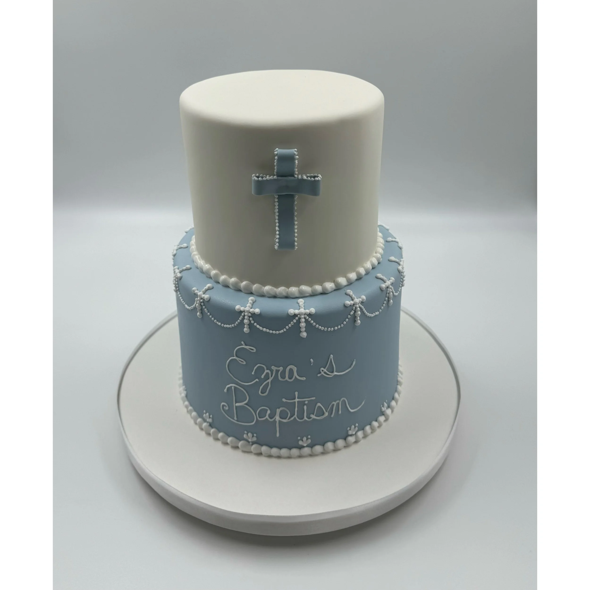 Two-tier baptism cake with white and blue fondant, decorated with pearl-like beads, a cross on the top tier.