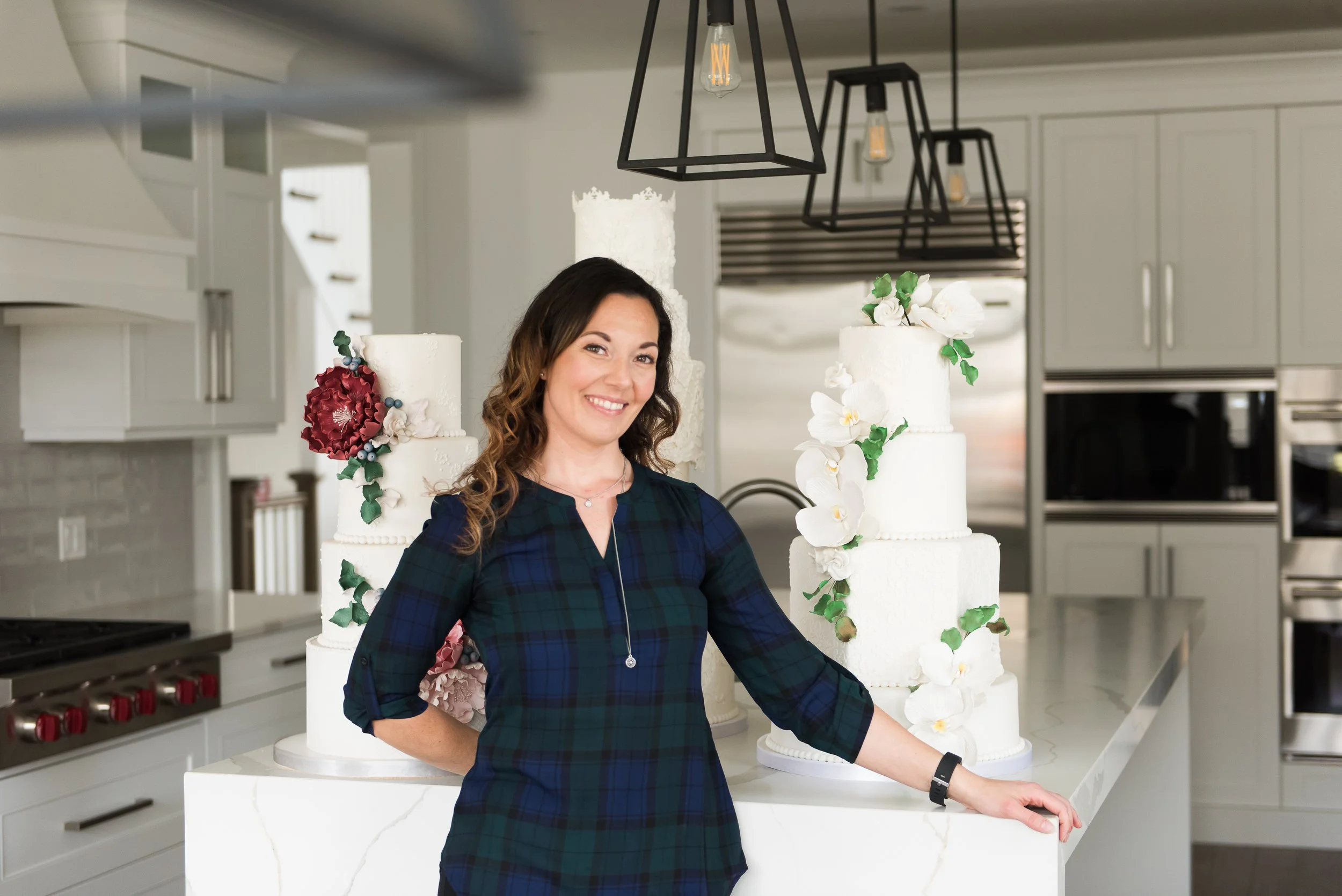 A woman with long, wavy brown hair and a blue plaid dress standing in a modern kitchen with two large white wedding cakes decorated with white flowers and green leaves behind her.