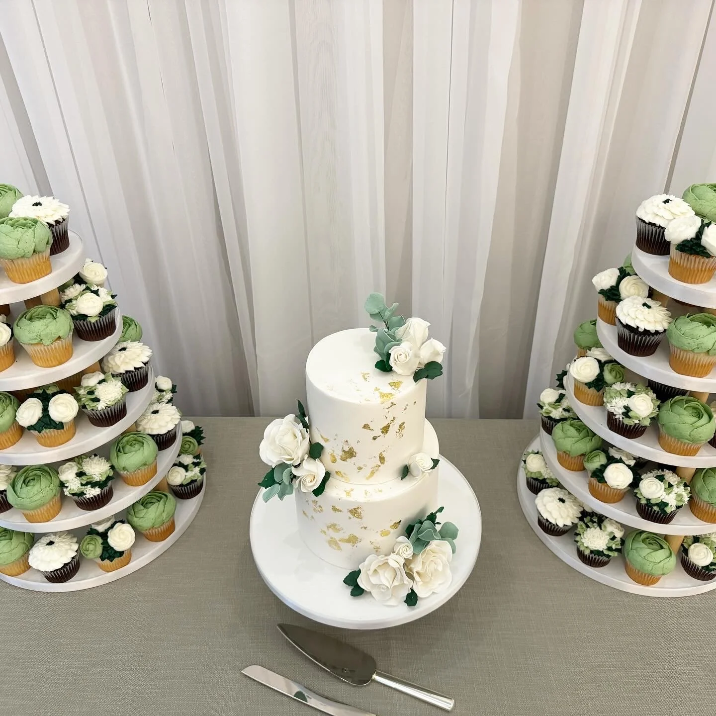 Two cupcake tiers with green, white, and brown cupcakes and a two-tier white wedding cake with white flowers and gold accents on a table.