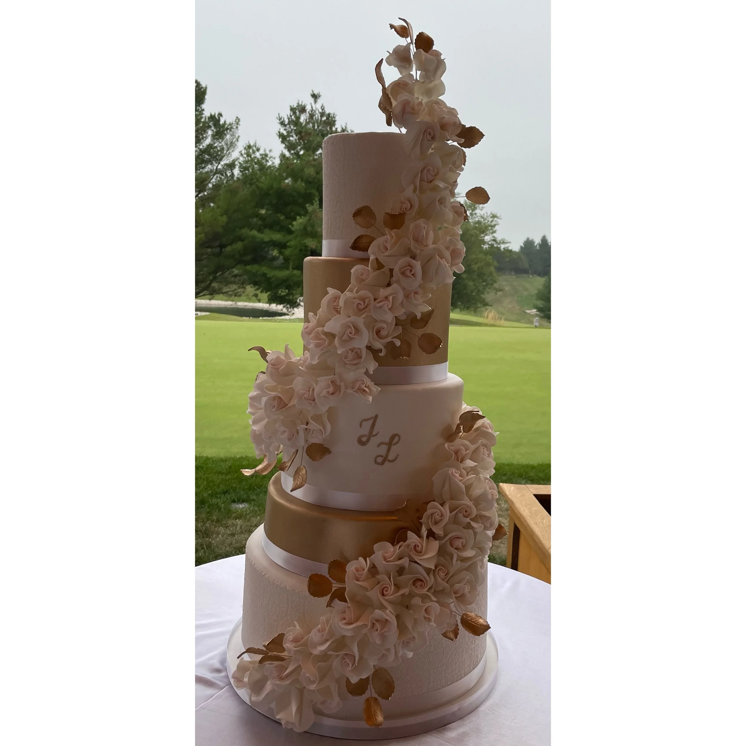 A four-tiered wedding cake decorated with white and gold accents, adorned with cascading white roses and gold-coloured leaves.